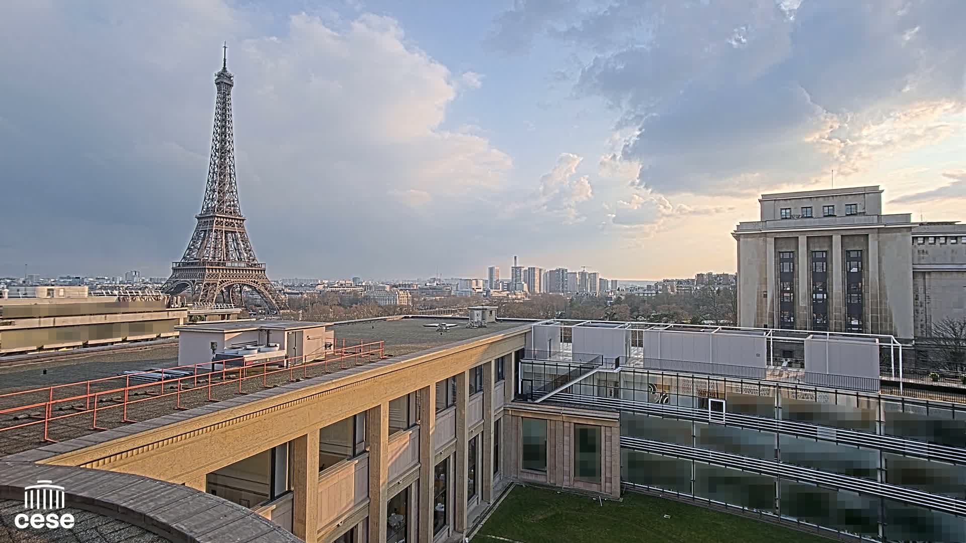 A Parisian cityscape under a partly cloudy sky, featuring the Eiffel Tower in the distance and a view from a rooftop overlooking other buildings.