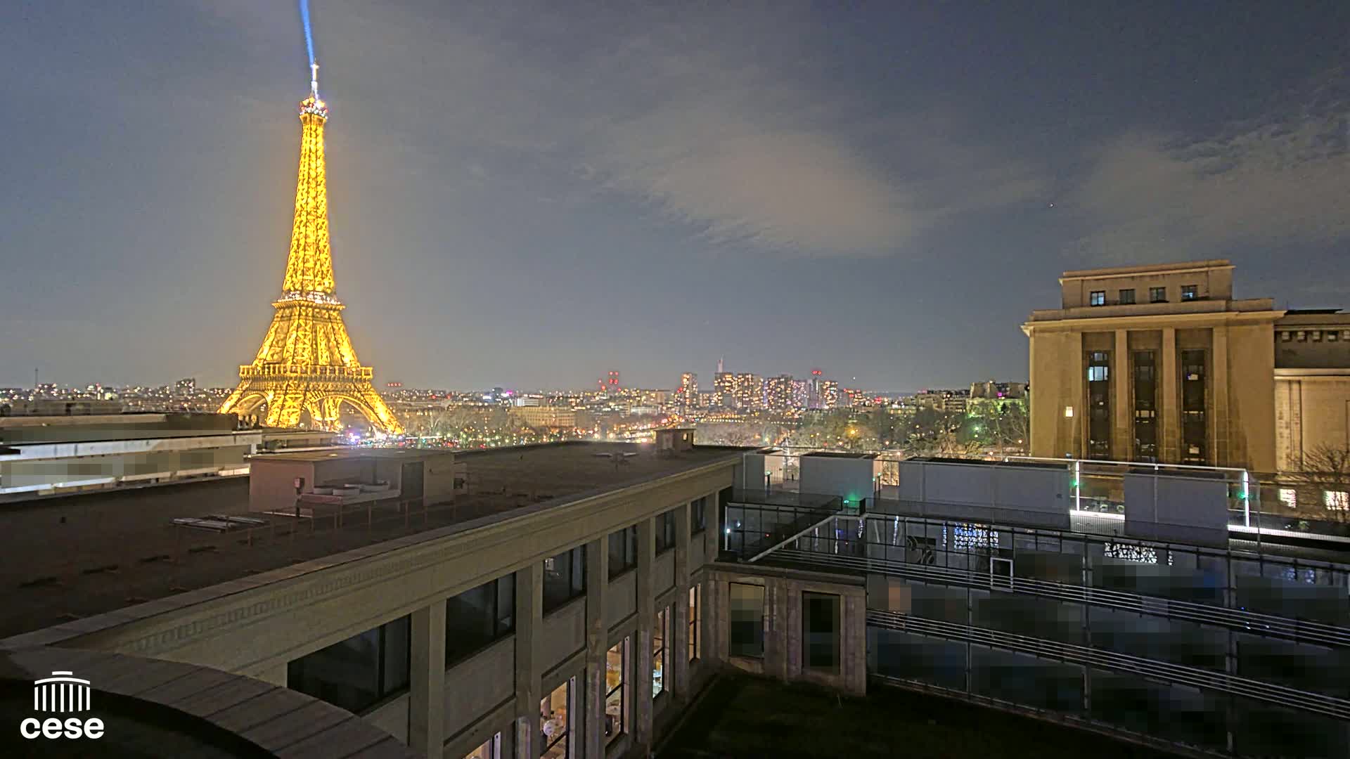 A brightly lit Eiffel Tower is visible at night from a rooftop overlooking a city, with a partly cloudy sky.