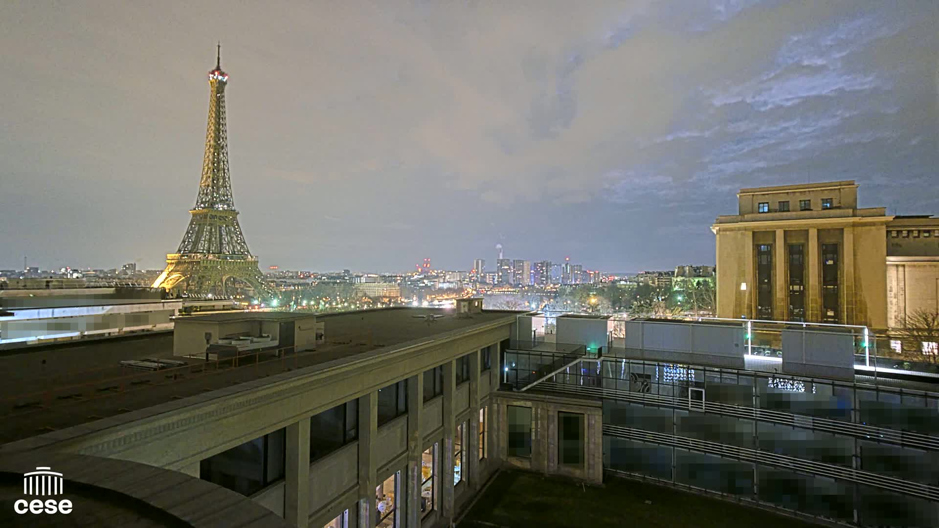 An illuminated Eiffel Tower is visible in the distance at night under a cloudy sky, from an elevated viewpoint overlooking city buildings and rooftops.