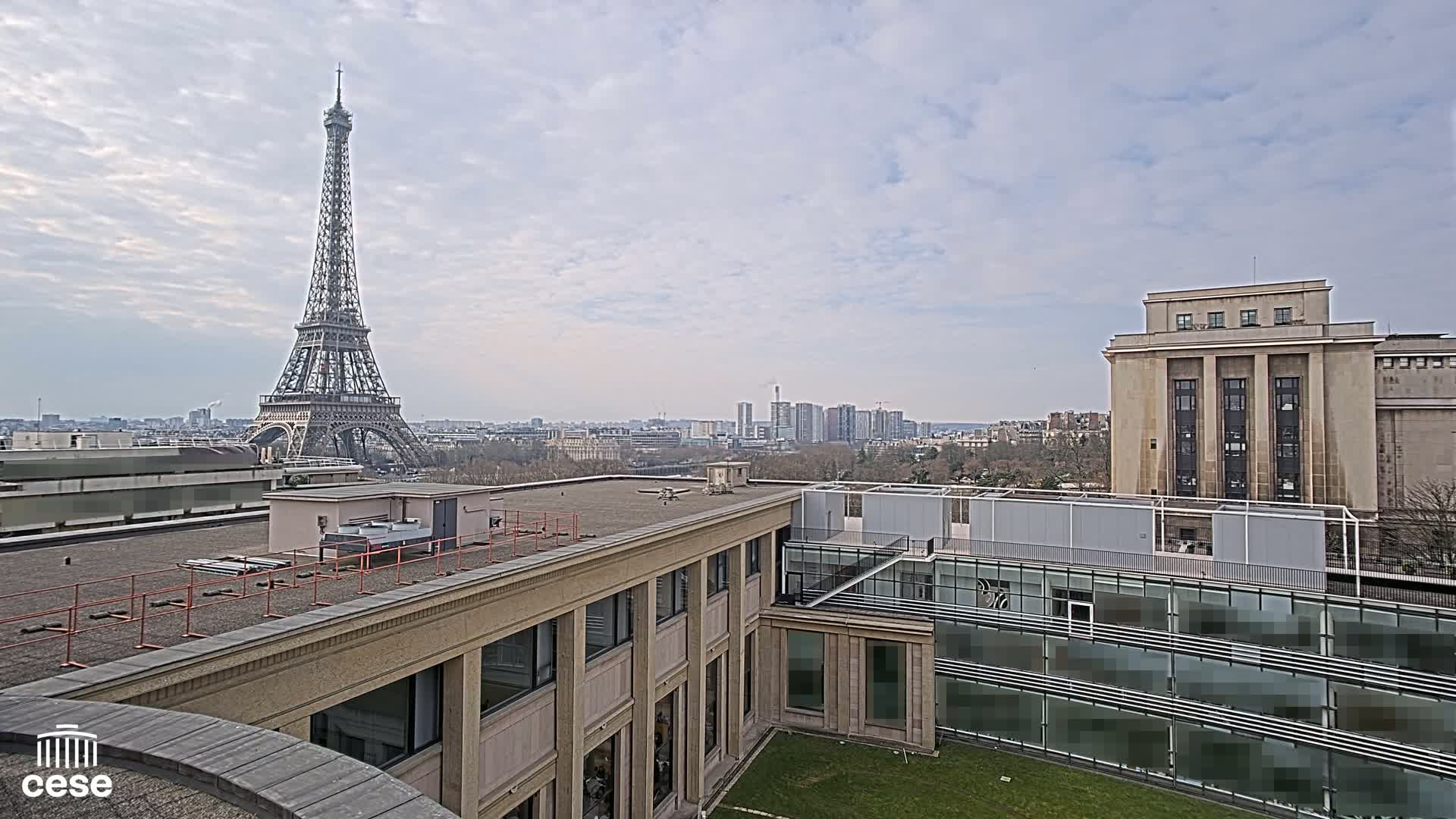 A rooftop view of the Eiffel Tower in the distance on a cloudy day, with modern and classical buildings in the foreground.