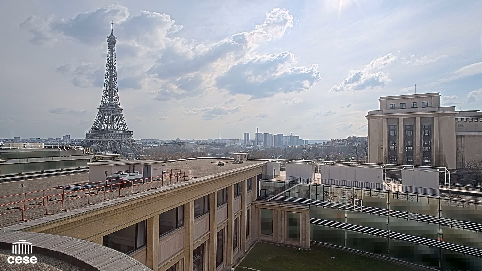 A view from a rooftop shows the Eiffel Tower in the distance under a partly cloudy sky, with a city skyline and other buildings nearby.