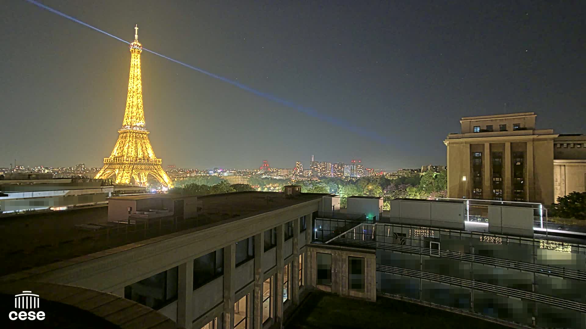 A nighttime view of the Eiffel Tower illuminated, city lights, and buildings from a rooftop vantage point.