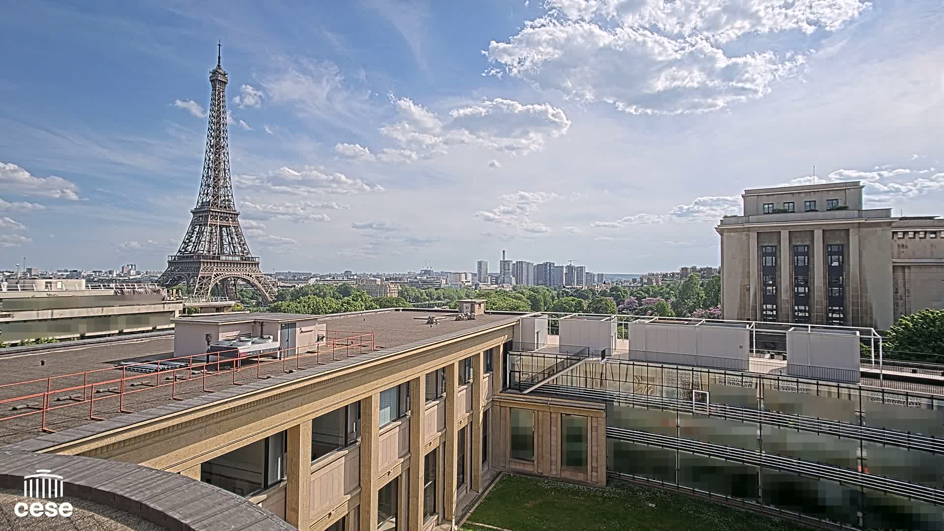 A rooftop view of Parisian buildings on a partly cloudy day, with the Eiffel Tower visible in the distance.