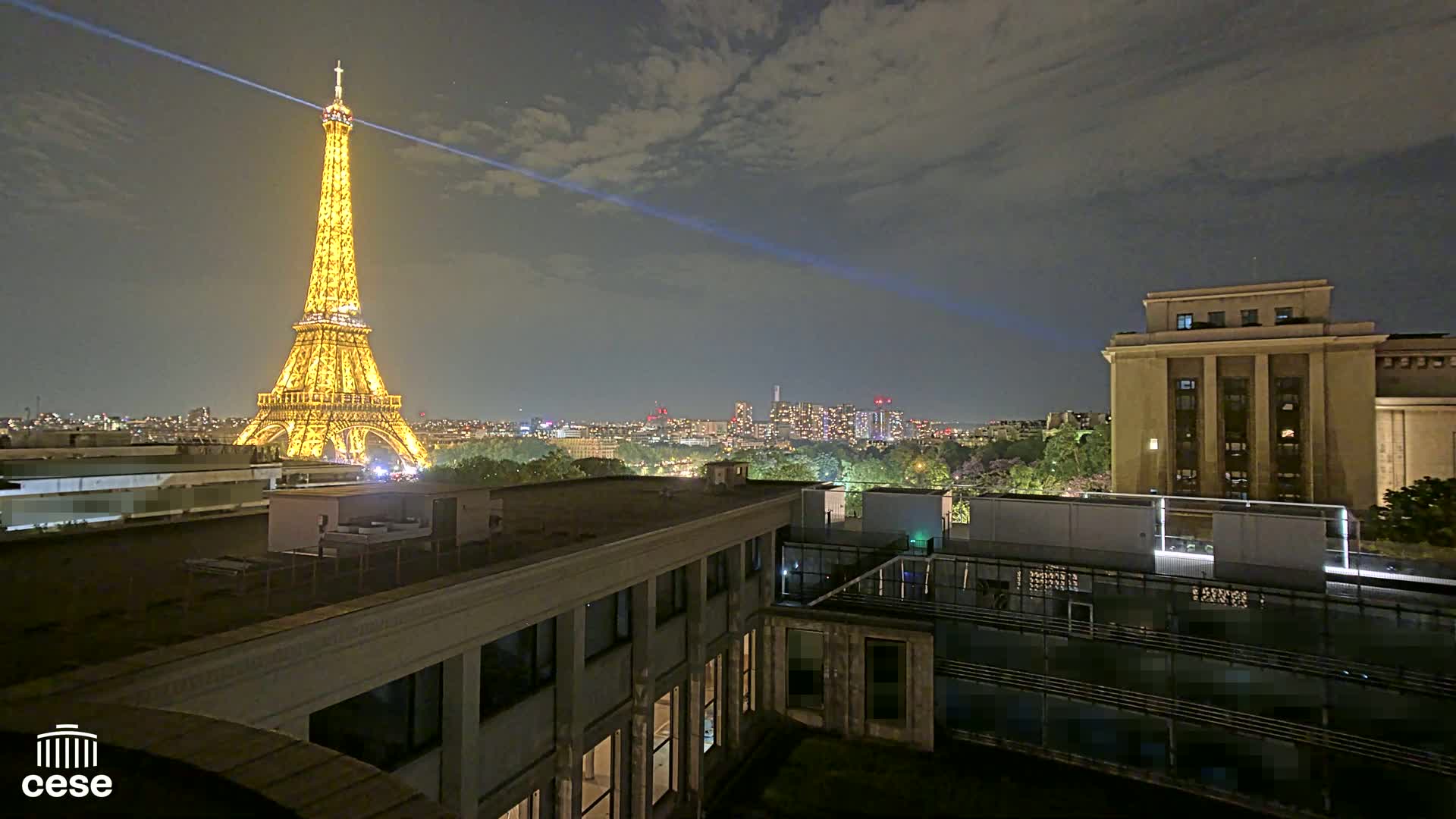 A nighttime view of the Eiffel Tower illuminated, a city skyline, and a building rooftop with a clear sky.
