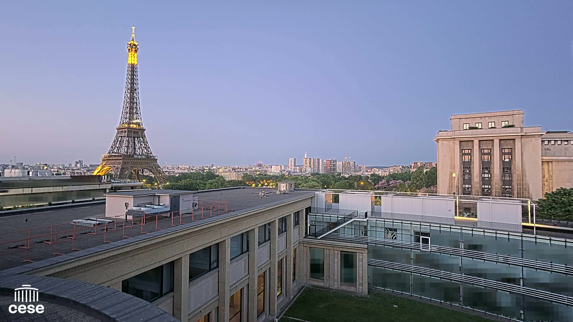 A view from a rooftop shows the Eiffel Tower illuminated at dusk, with a cityscape extending to the horizon and modern buildings in the immediate foreground.