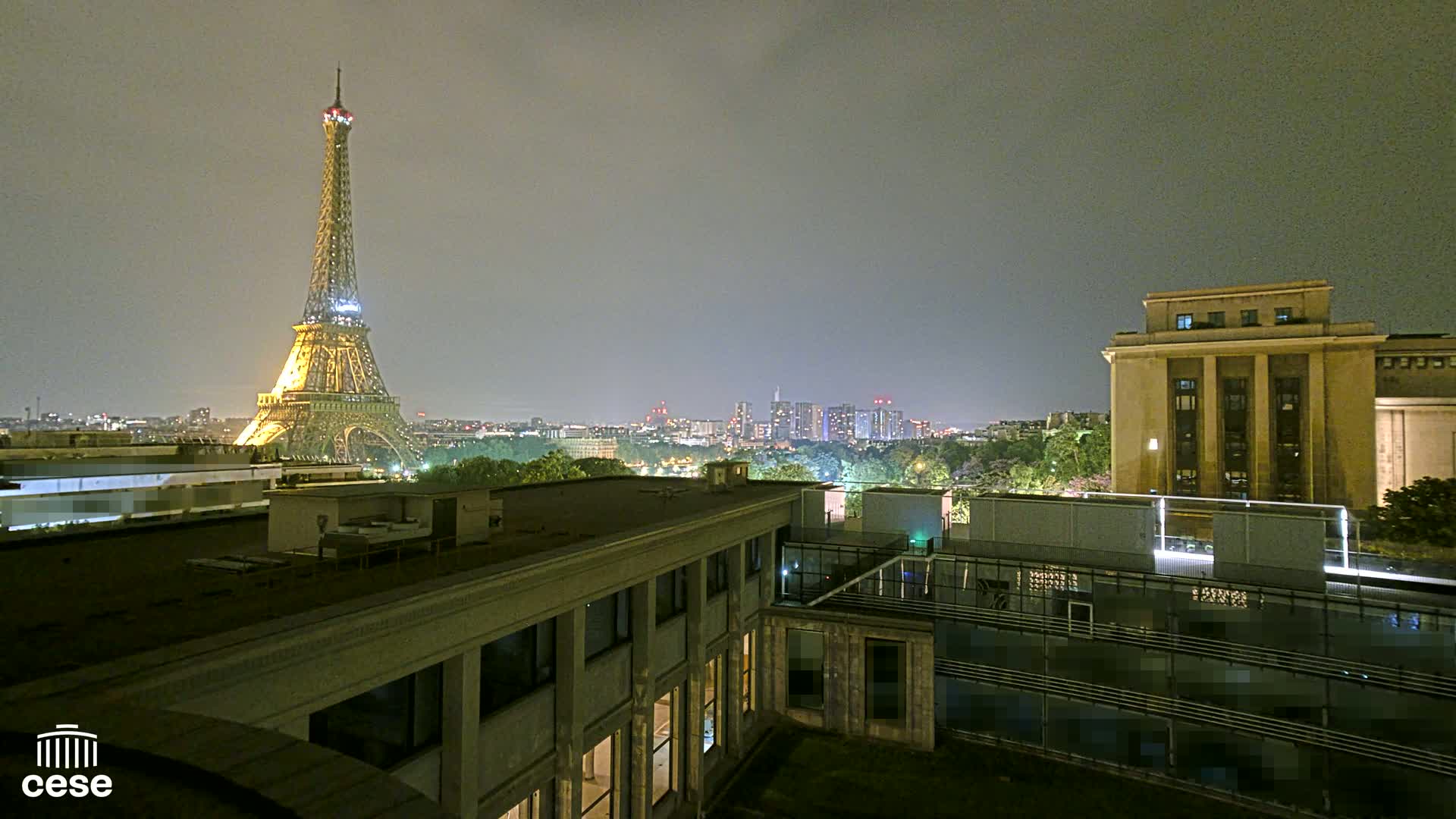 A nighttime view of the Eiffel Tower illuminated against a dusky sky, from a rooftop overlooking a city with a large building in the foreground.