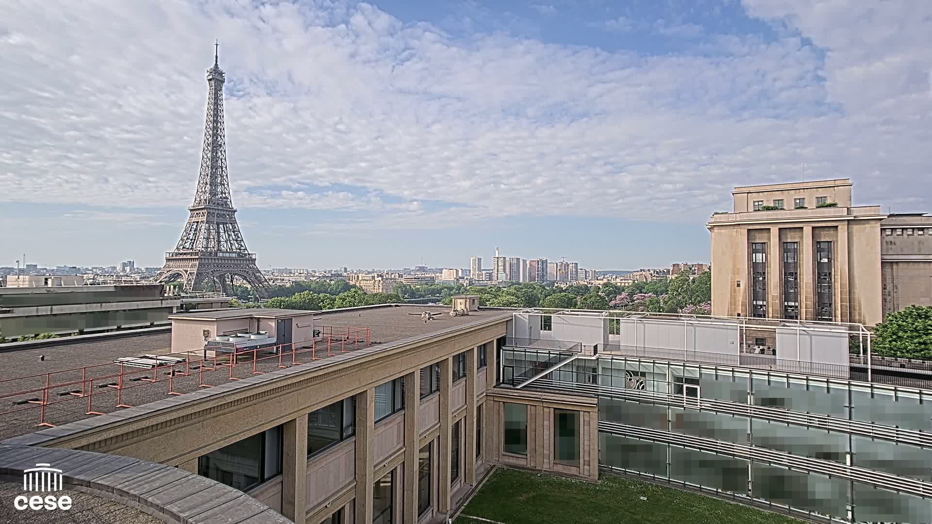 A rooftop view of Paris on a partly cloudy day shows the Eiffel Tower in the distance, alongside city buildings and green trees.