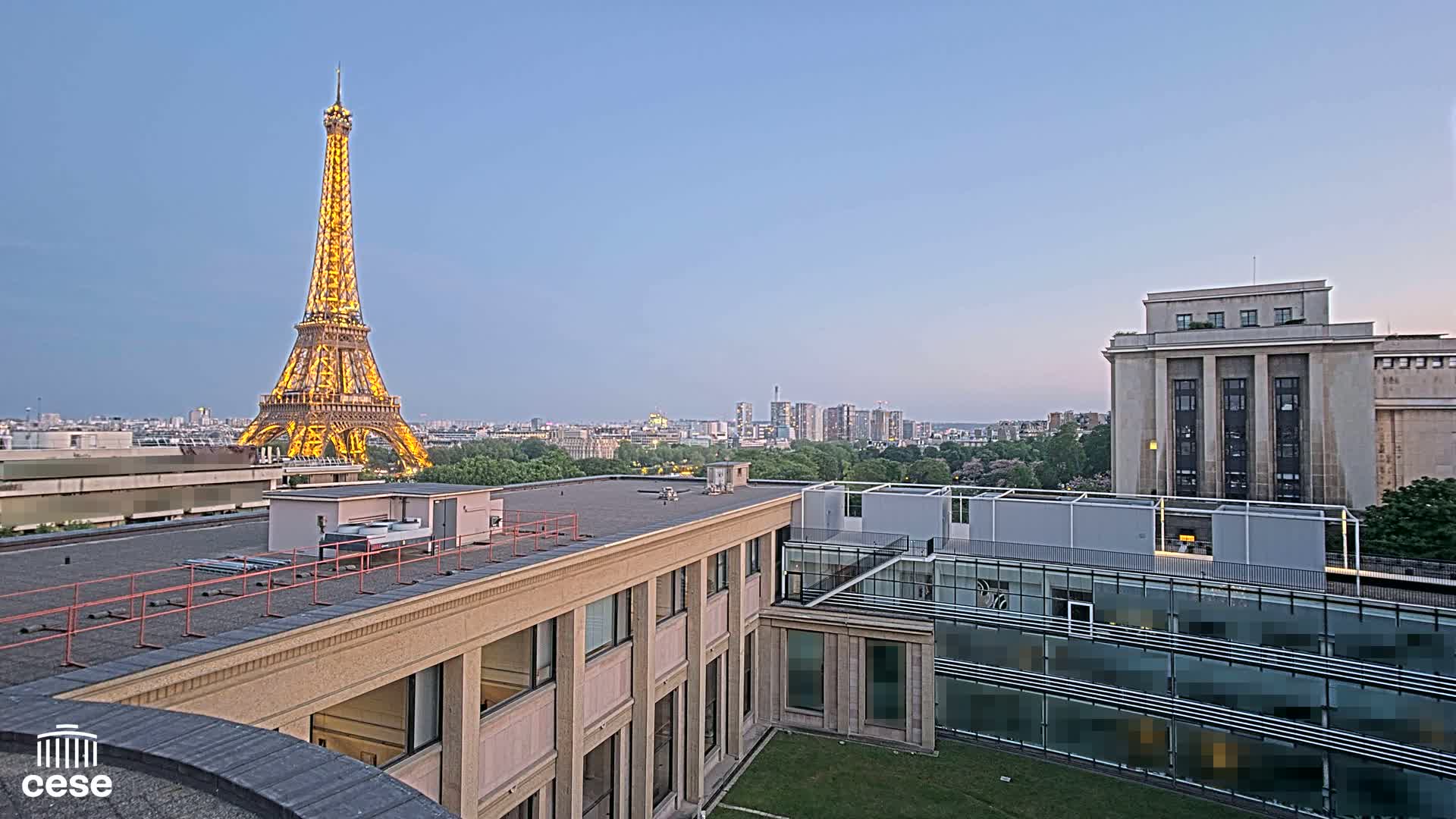 A brightly lit Eiffel Tower is visible in the distance from a rooftop vantage point overlooking a city under a clear, twilight sky.