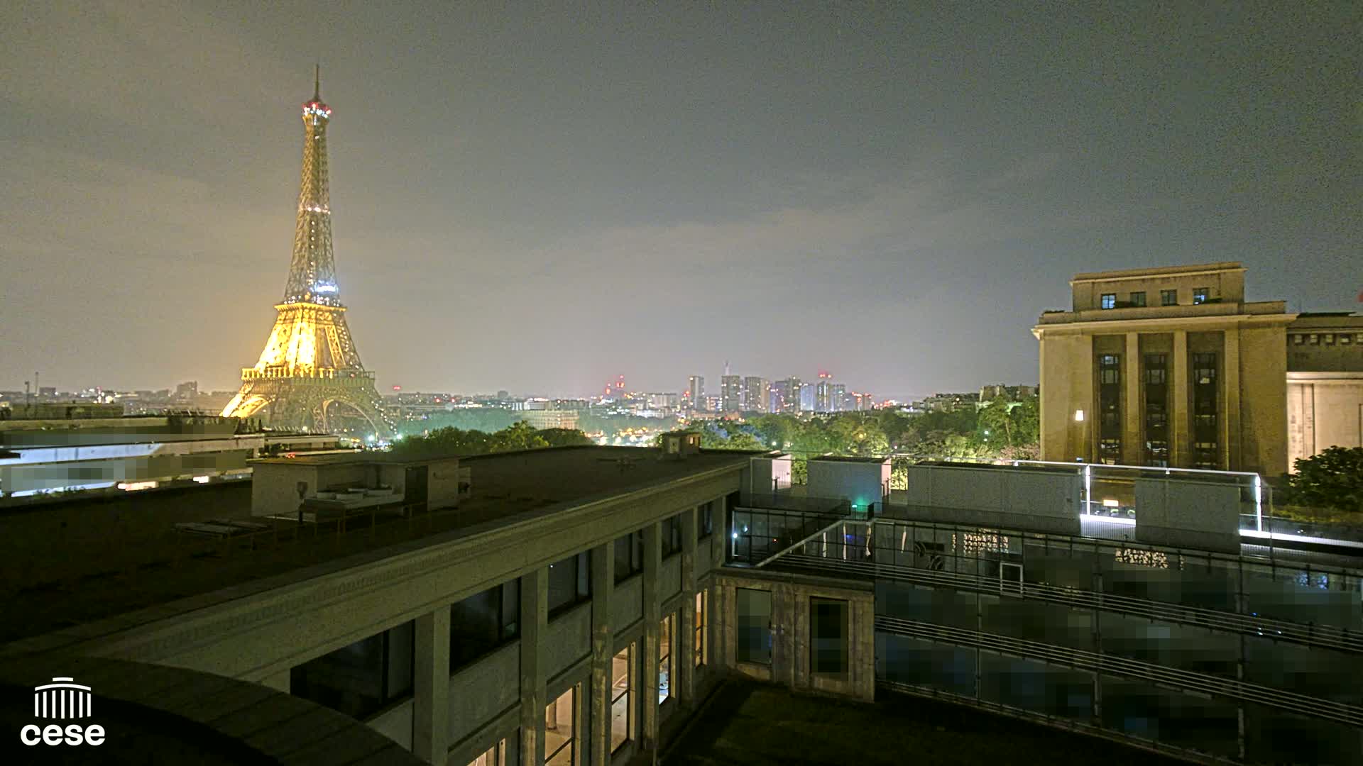 The Eiffel Tower is illuminated at night, viewed from a rooftop overlooking a city skyline and other buildings.