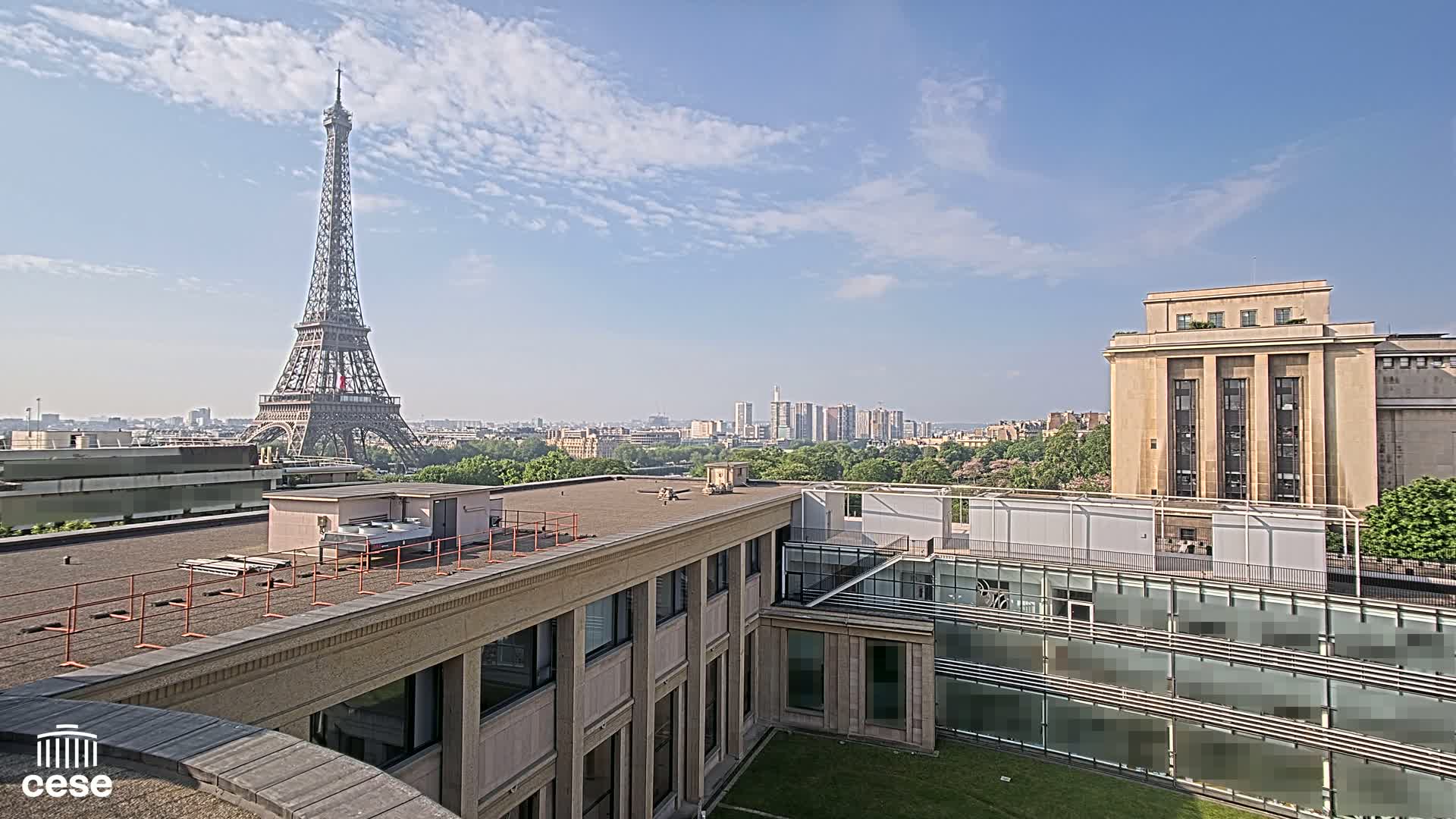 A rooftop view of Paris on a partly cloudy day shows the Eiffel Tower in the distance, with city buildings and green trees in the foreground.