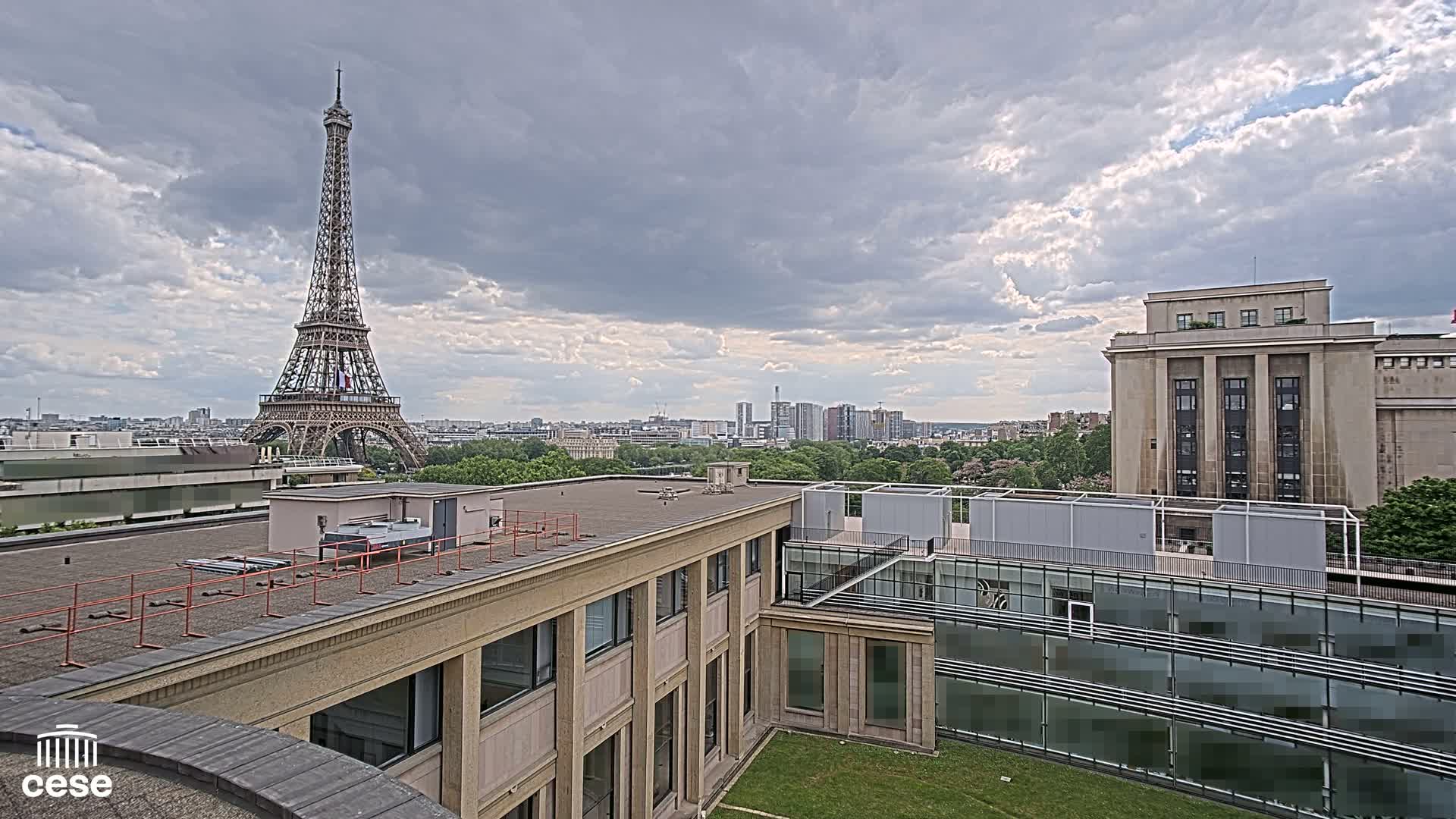 A view from a rooftop shows the Eiffel Tower in the distance under a cloudy sky, with other buildings and green spaces in the foreground.