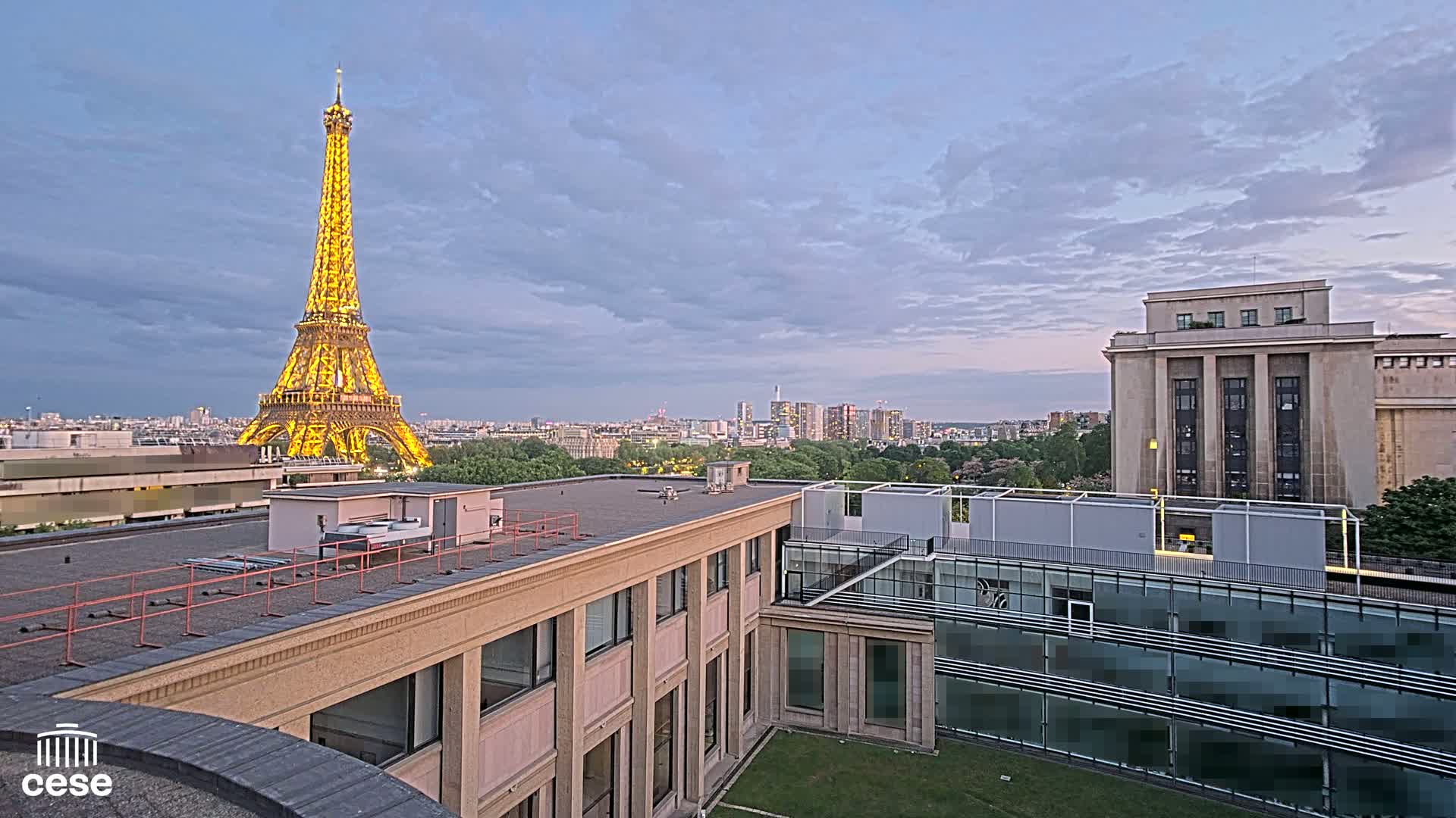 A rooftop view of the Eiffel Tower illuminated at dusk, overlooking a cityscape with a large building in the foreground.