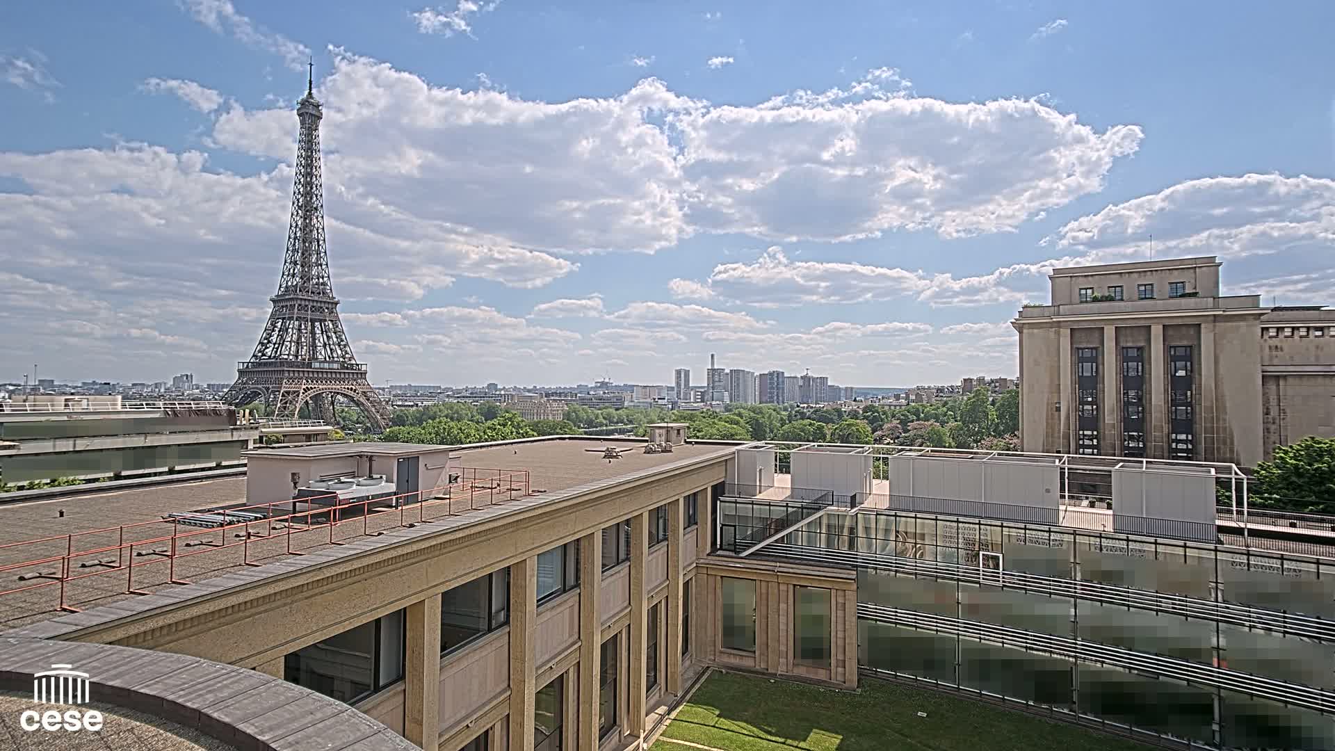 A rooftop view of Parisian buildings, including the Eiffel Tower in the distance, under a partly cloudy sky.