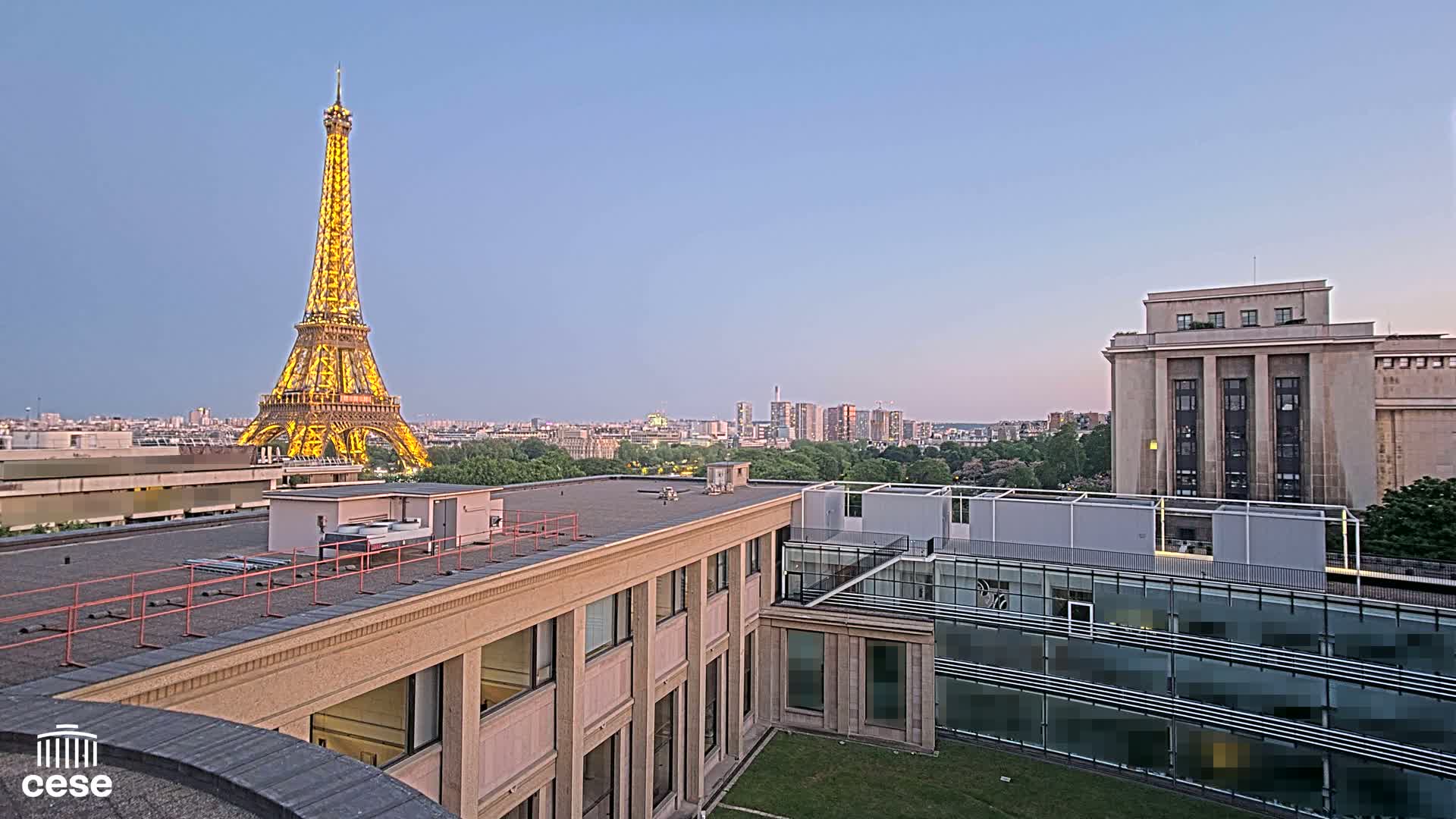 A rooftop view of Paris at dusk shows the Eiffel Tower illuminated, city buildings, and green spaces.