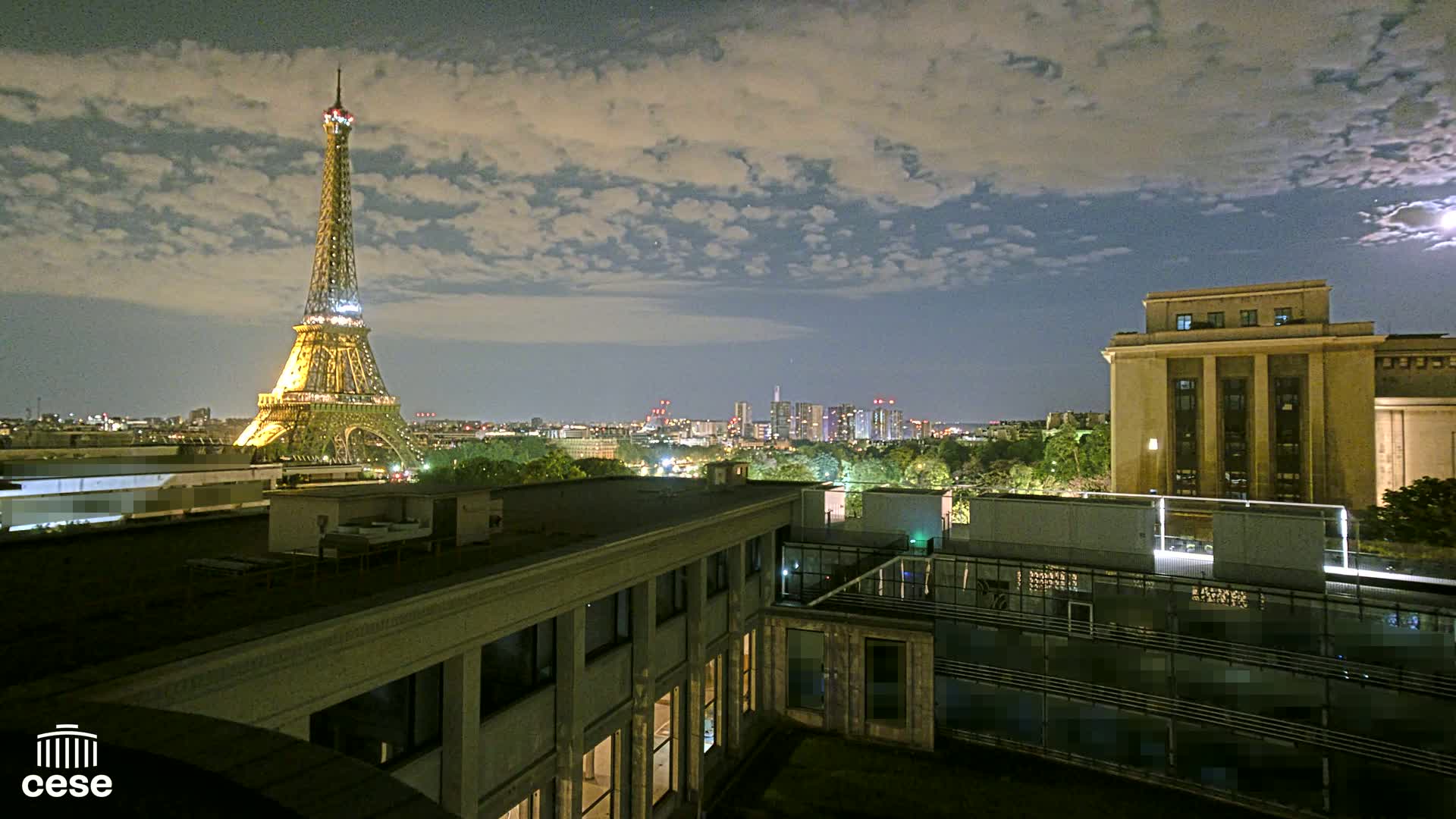 A nighttime view of the Eiffel Tower and a cityscape, under a partly cloudy sky, from a rooftop vantage point.