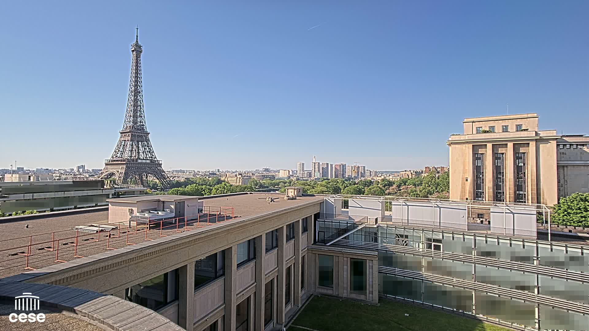 A rooftop view on a clear, sunny day shows the Eiffel Tower in the distance, beyond a cityscape, and several modern buildings.