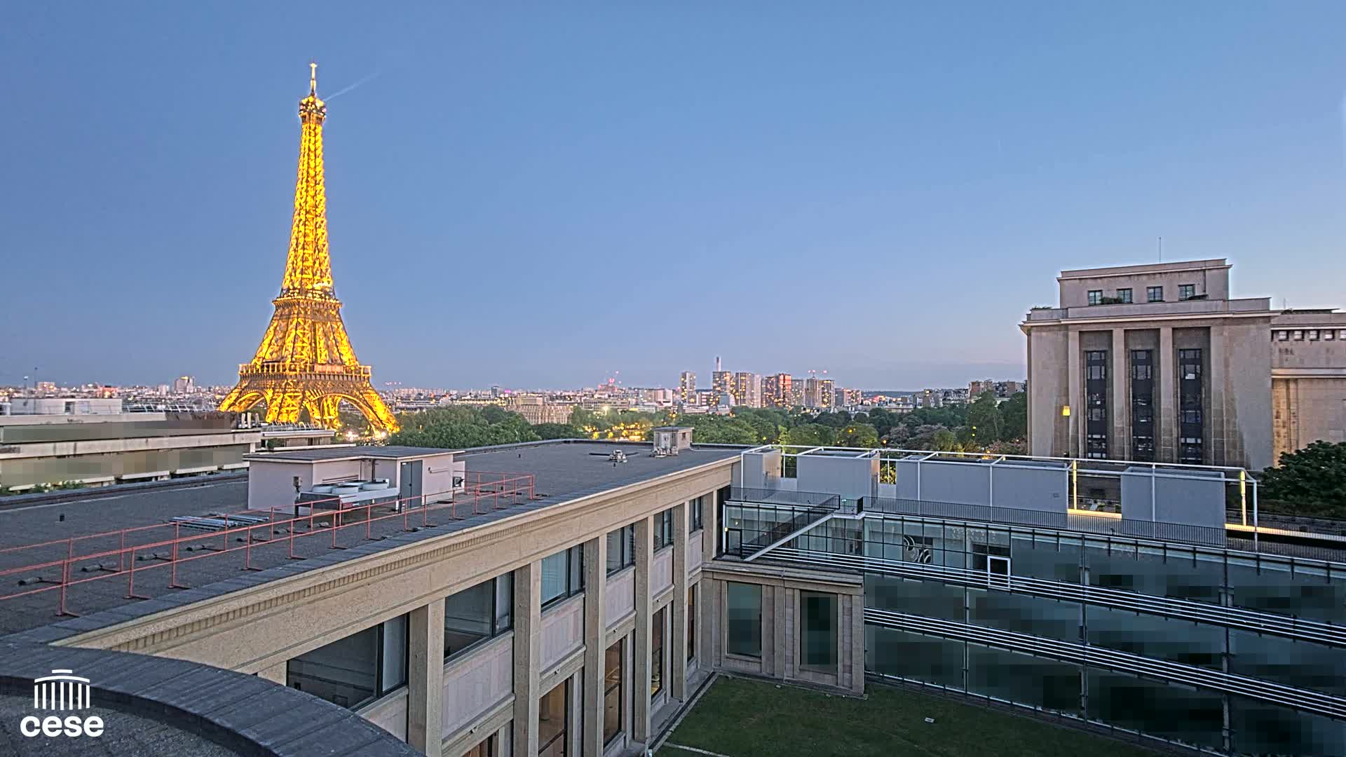 A brightly lit Eiffel Tower is visible in the distance from a rooftop vantage point overlooking a city at dusk.