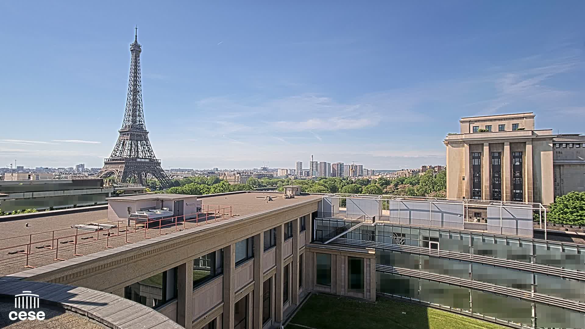 A rooftop view of Paris on a sunny day shows the Eiffel Tower in the distance, city buildings, and green trees.