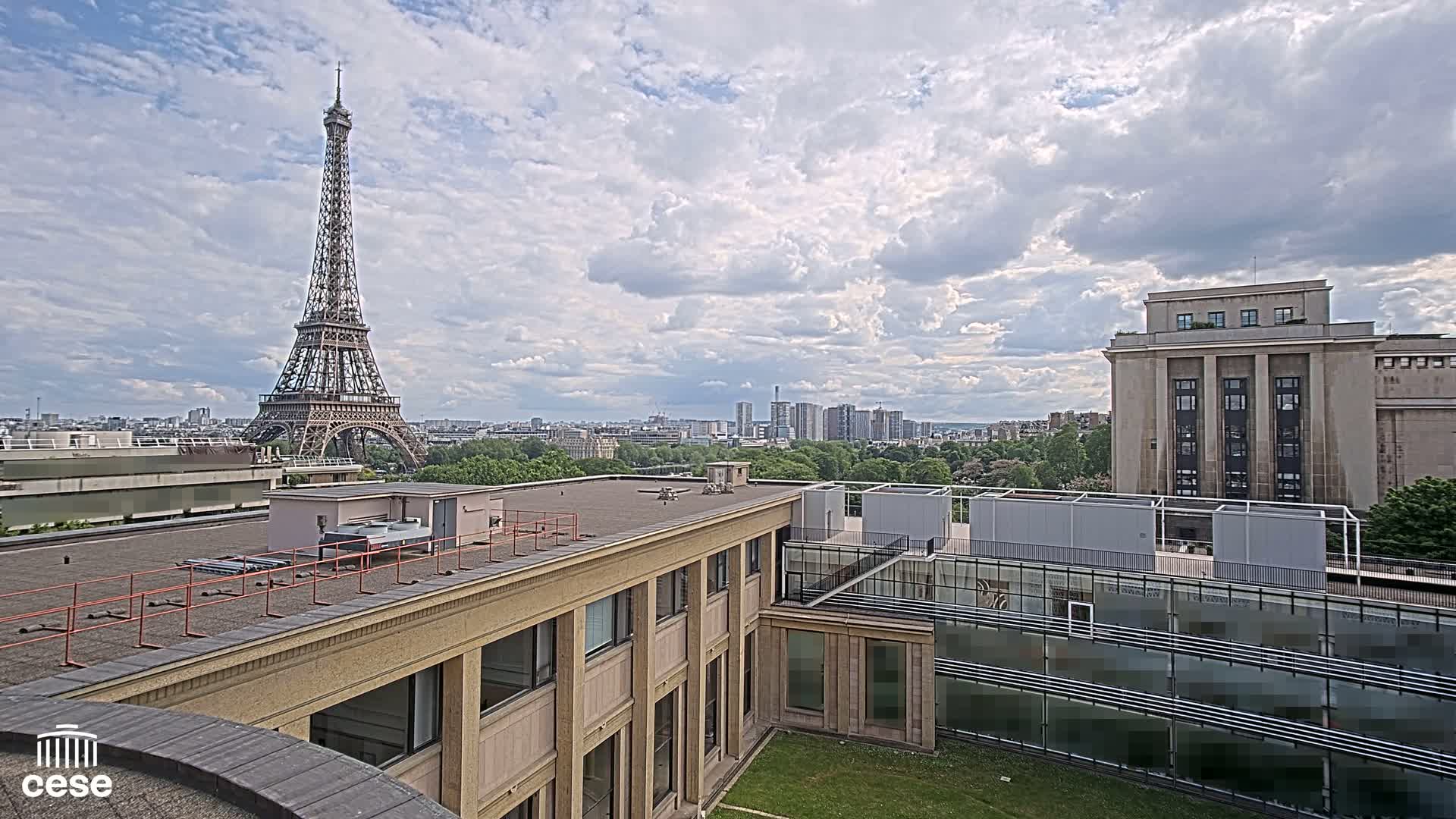 A view from a rooftop shows the Eiffel Tower in the distance under a partly cloudy sky, with other buildings and green spaces in the foreground.