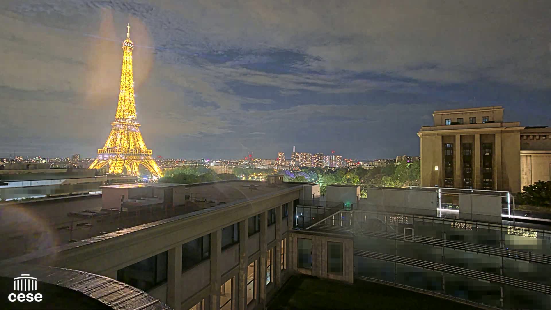 A nighttime view of the Eiffel Tower illuminated, across a cityscape under a partly cloudy sky, from a rooftop vantage point.