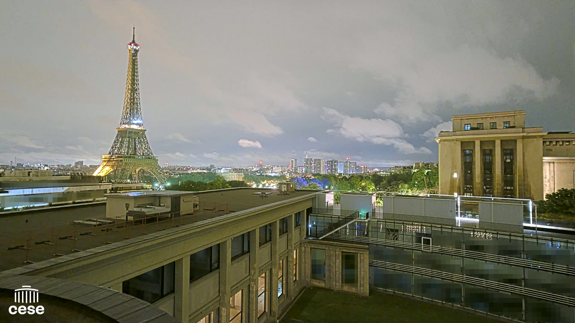 A nighttime cityscape view of the Eiffel Tower and a portion of Paris, under a cloudy sky, as seen from a rooftop.