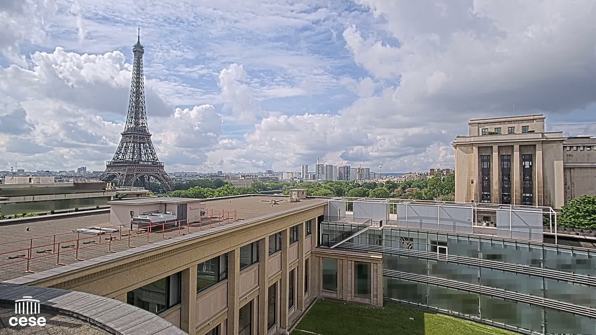 A Parisian cityscape under a partly cloudy sky features the Eiffel Tower in the distance, viewed from atop a building with a flat roof and modern architecture.