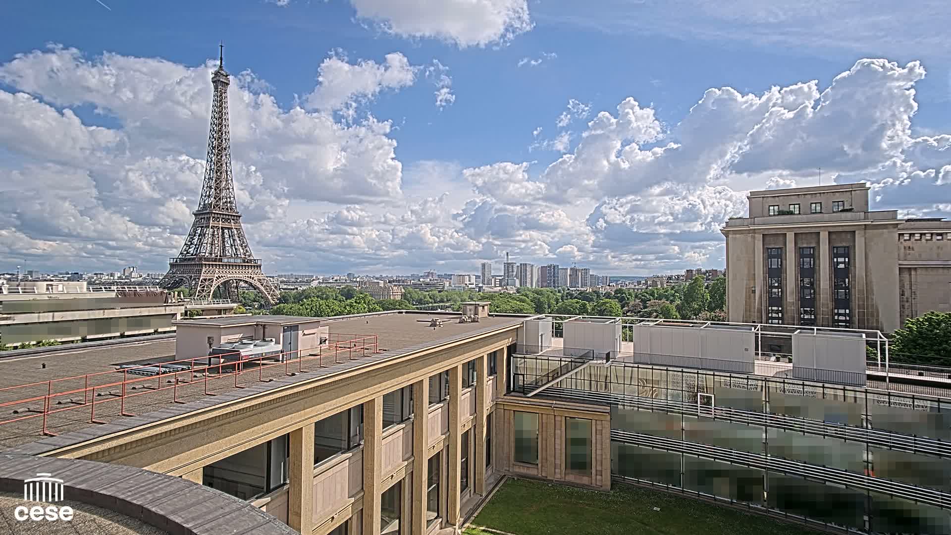 A rooftop view shows the Eiffel Tower in the distance under a partly cloudy sky, with a city skyline and a large building in the foreground.