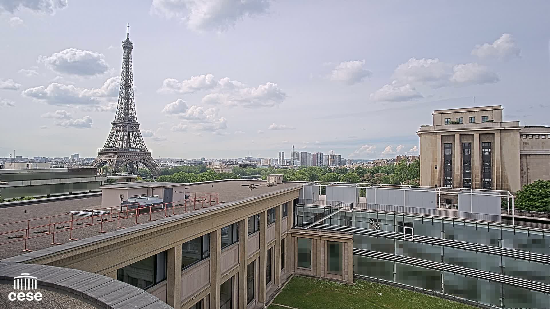 The Eiffel Tower is visible in the distance under a partly cloudy sky, from a rooftop vantage point overlooking other buildings and a cityscape.