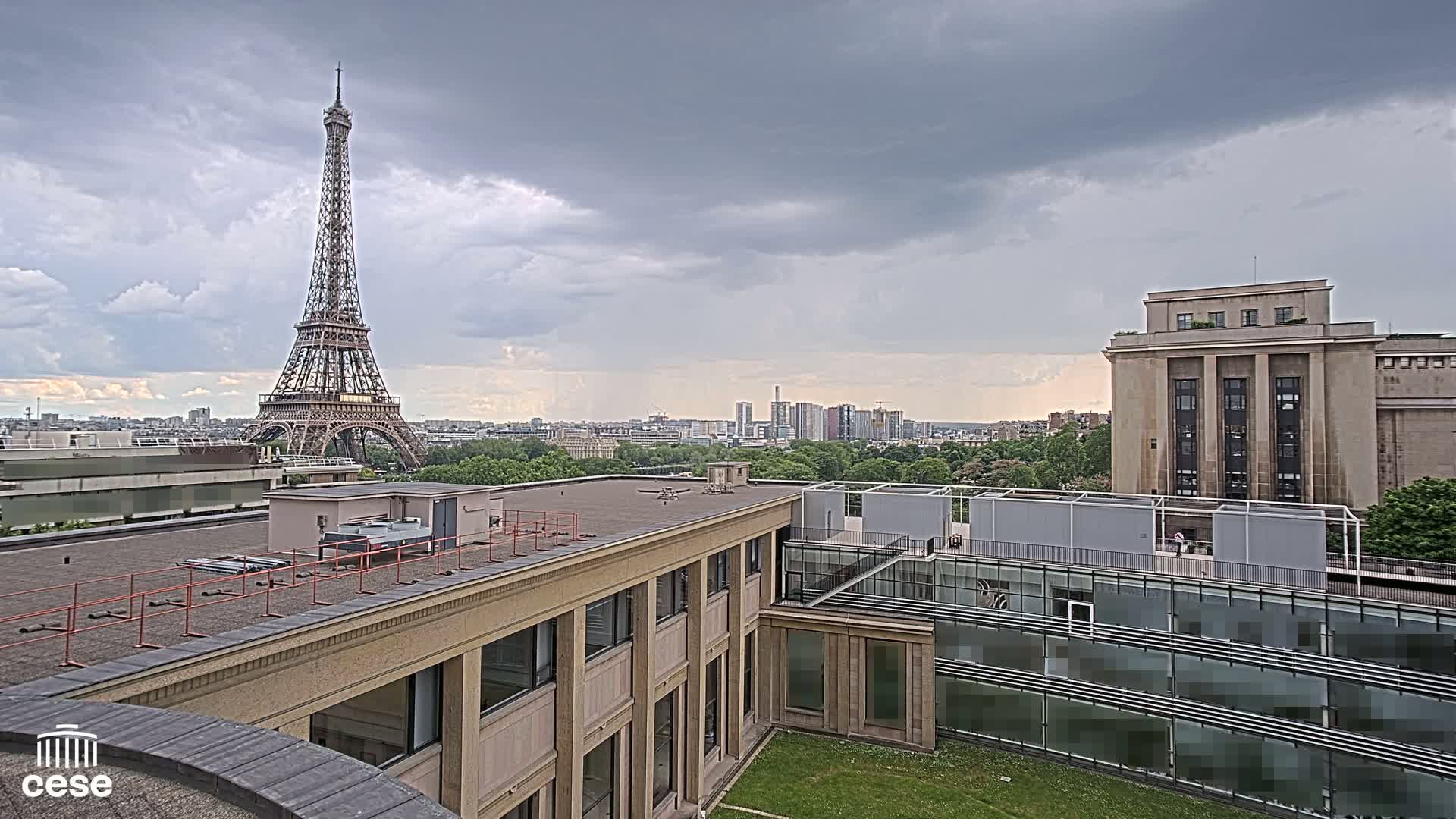 A rooftop view of Parisian buildings under a cloudy sky, with the Eiffel Tower visible in the distance.