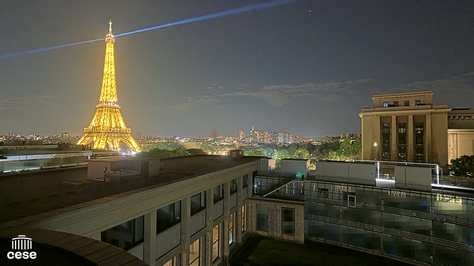 A nighttime view of the Eiffel Tower illuminated, with city lights in the distance and a building's rooftop in the foreground under a clear, dark sky.