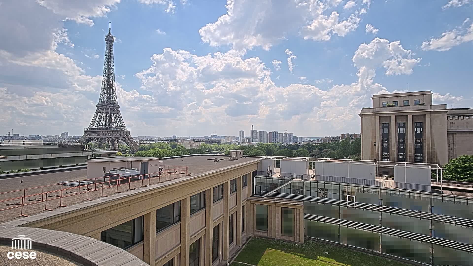 A partly cloudy sky overlooks a Parisian cityscape, including the Eiffel Tower, from a rooftop with tan buildings and a small grassy courtyard.