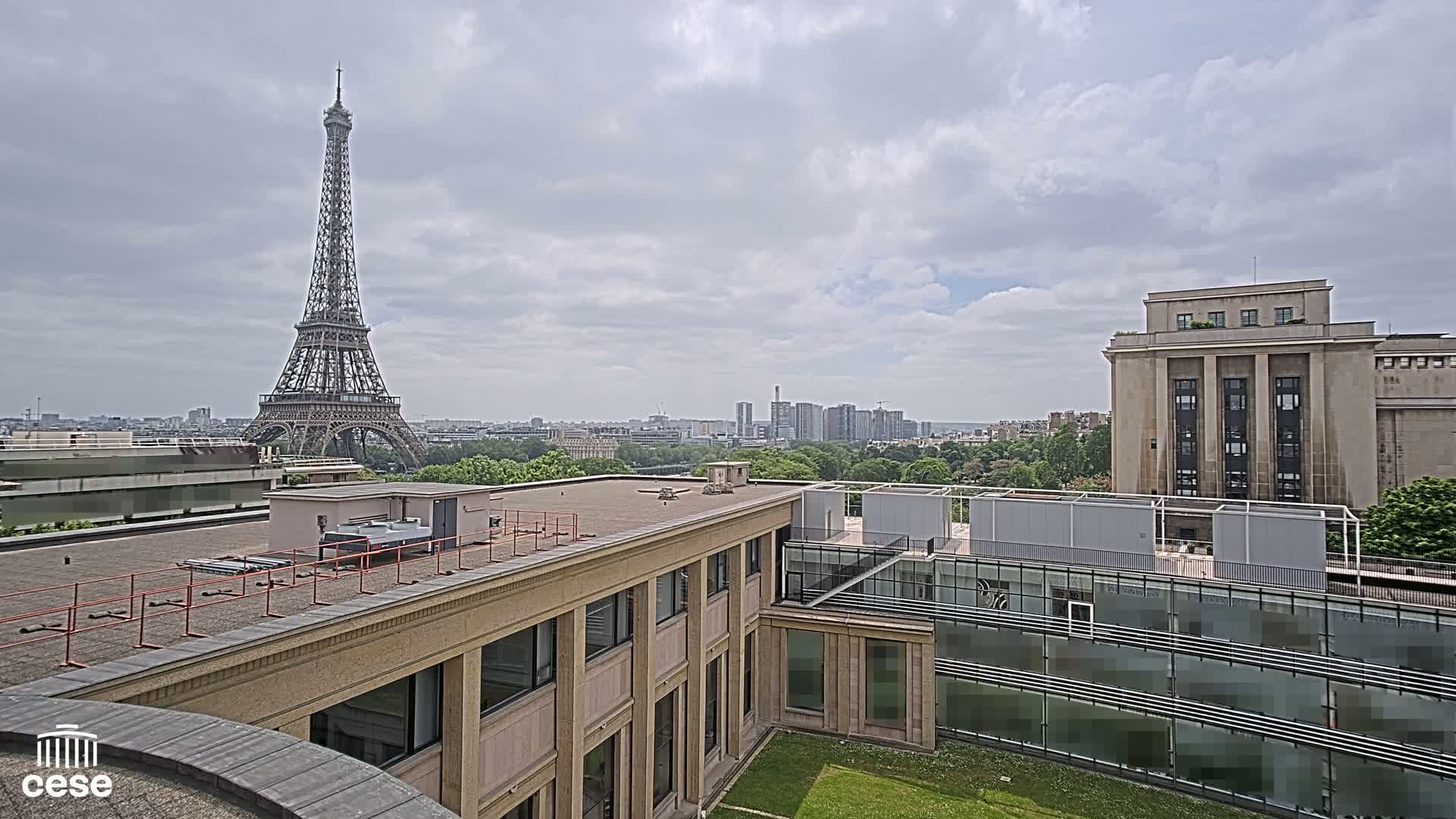 A view from a rooftop shows the Eiffel Tower in the distance under a cloudy sky, with other buildings and green spaces in the foreground.