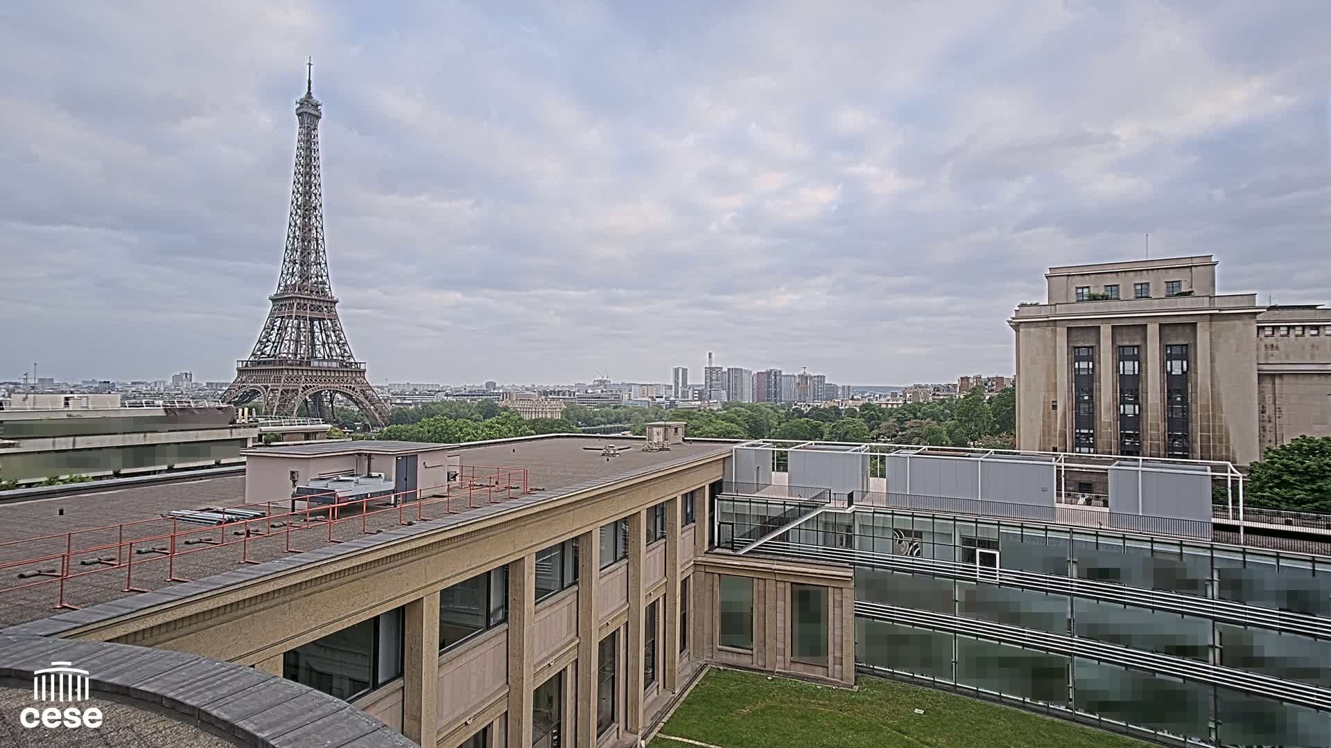 A rooftop view shows the Eiffel Tower in the distance under a cloudy sky, with several buildings and green spaces in the foreground.