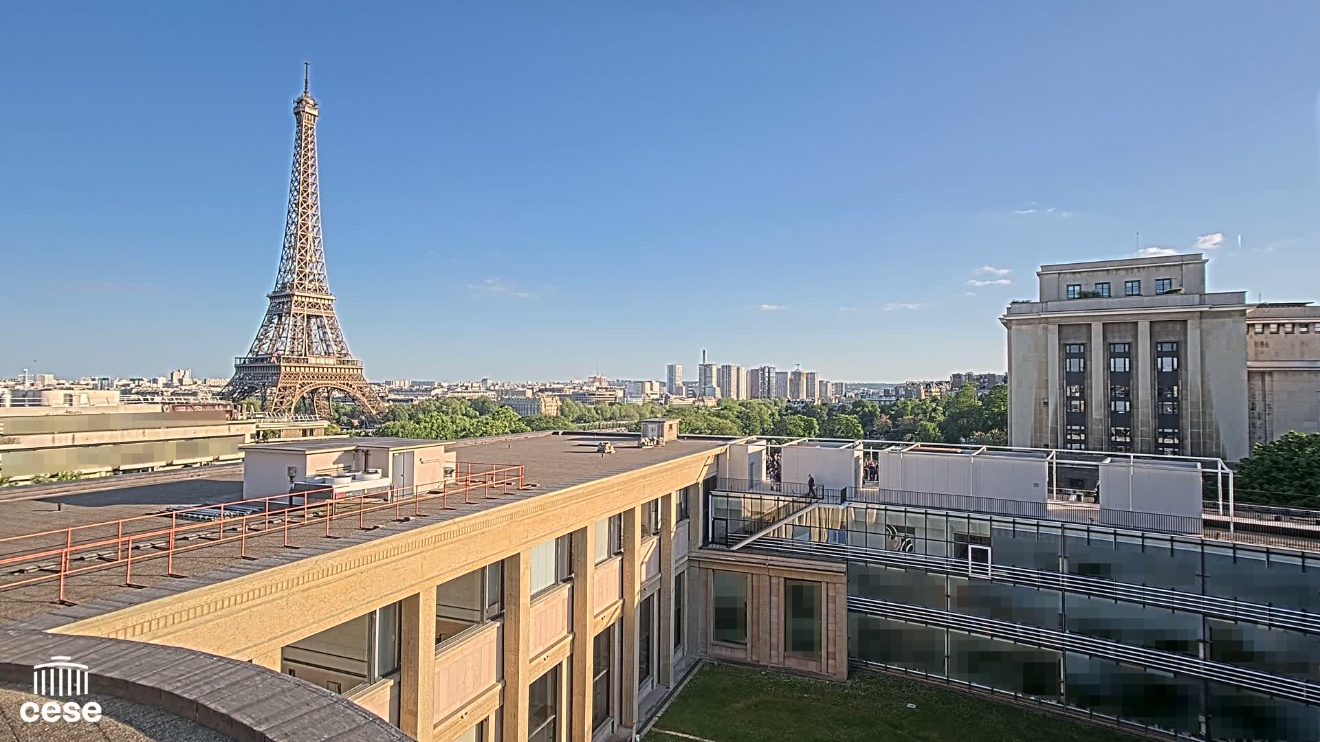 A rooftop view shows the Eiffel Tower in the distance under a clear blue sky, overlooking a city skyline and a modern building with a courtyard.