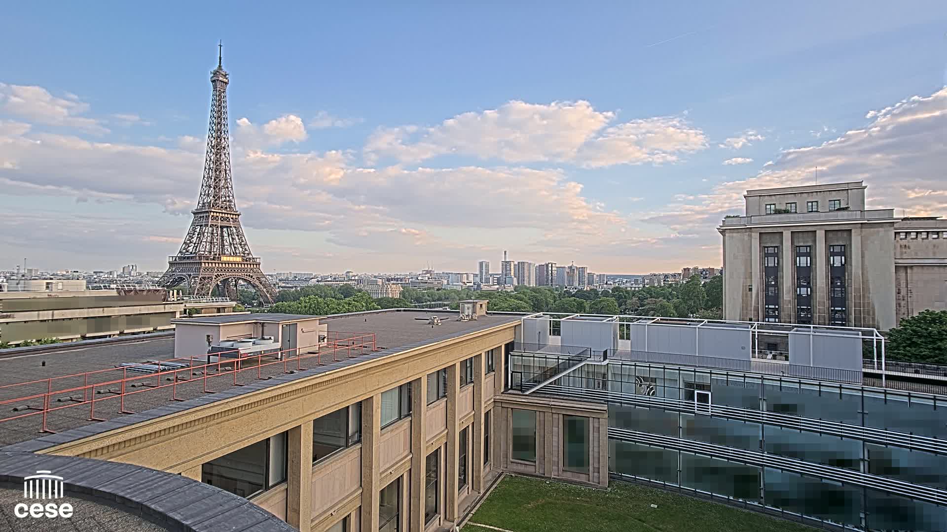 A rooftop view shows the Eiffel Tower in the distance under a partly cloudy sky, with city buildings and greenery in the foreground.