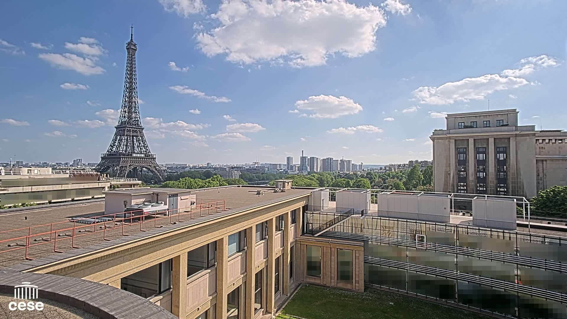 A rooftop view of Paris on a partly sunny day shows the Eiffel Tower in the distance,  a large building in the foreground, and a cityscape beyond.