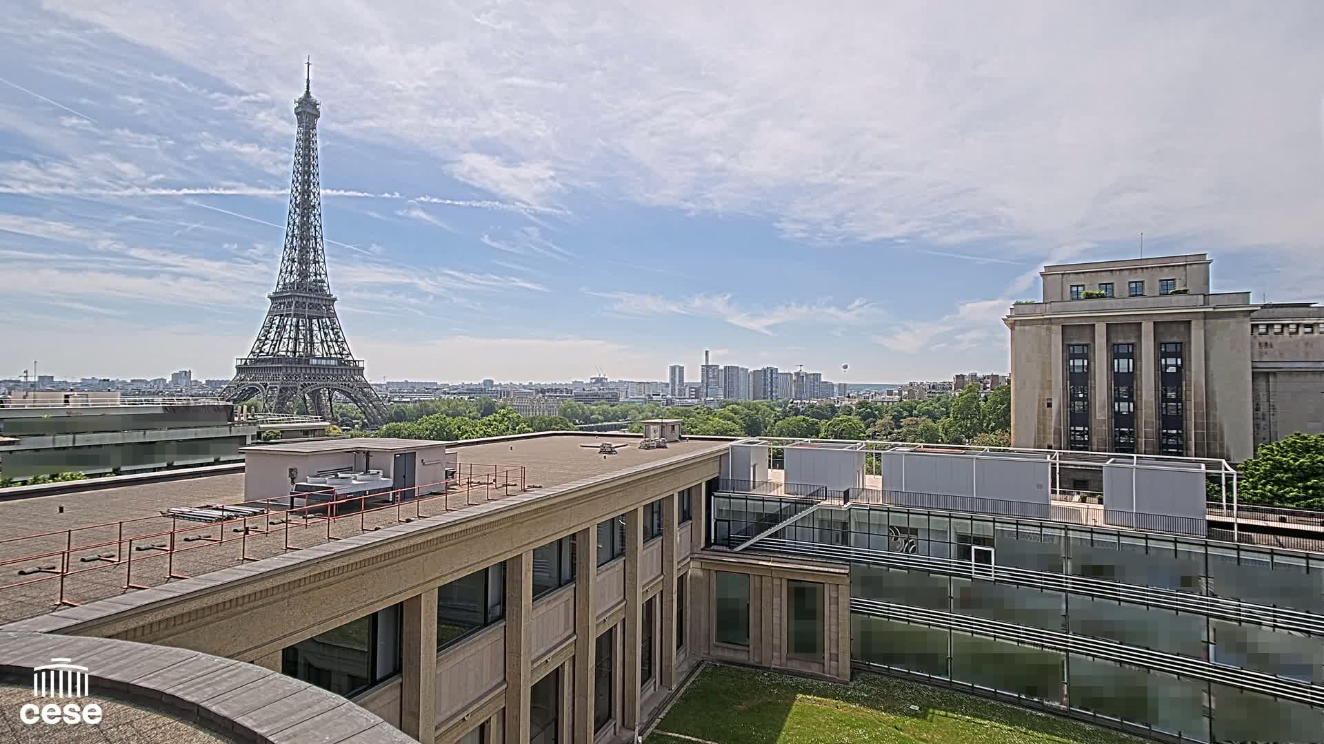 A rooftop view of Parisian buildings, including the Eiffel Tower in the distance, under a partly cloudy sky.