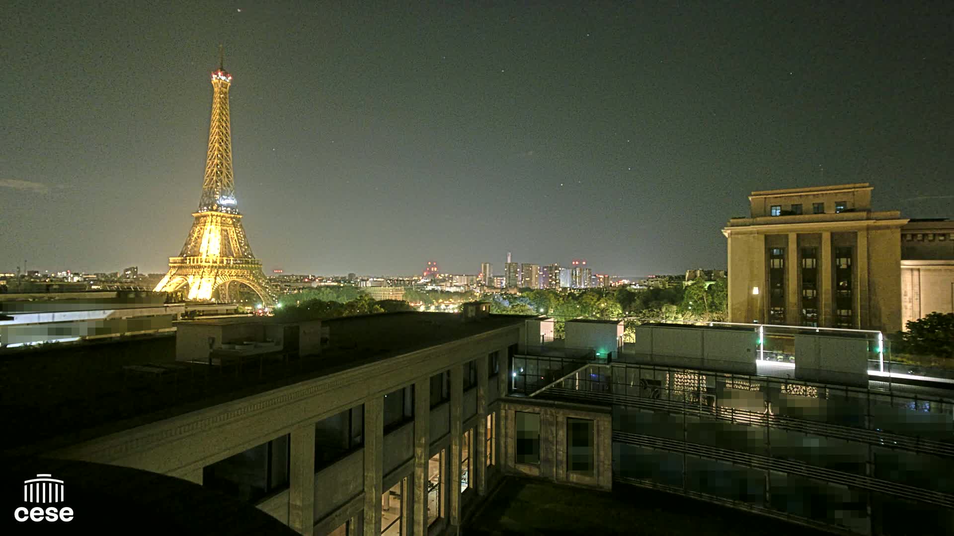 A nighttime view of the Eiffel Tower illuminated against a cityscape, seen from a rooftop overlooking other buildings under a clear, dark sky.