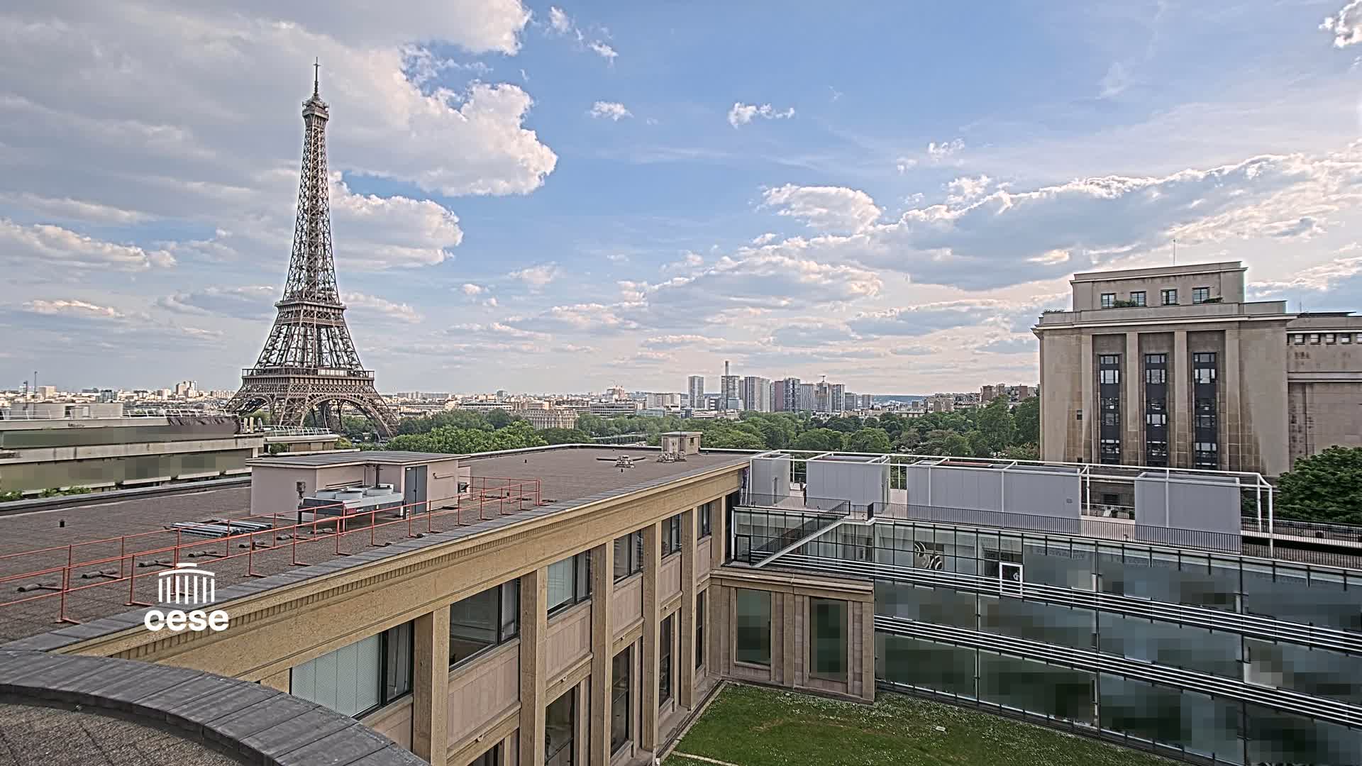 A Parisian rooftop view shows the Eiffel Tower in the distance under a partly cloudy sky, with other buildings and greenery in the foreground.