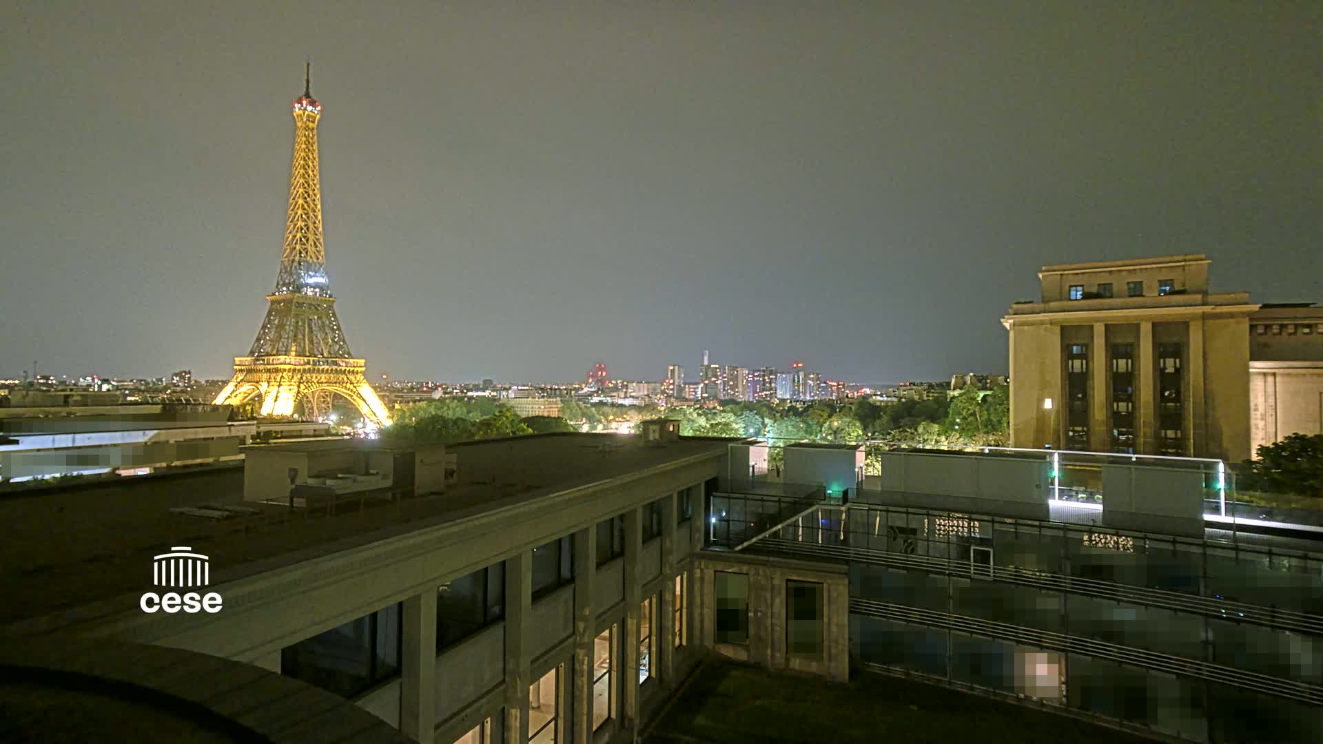 A nighttime cityscape featuring the Eiffel Tower illuminated, alongside a large building, and a city skyline under a cloudy sky.