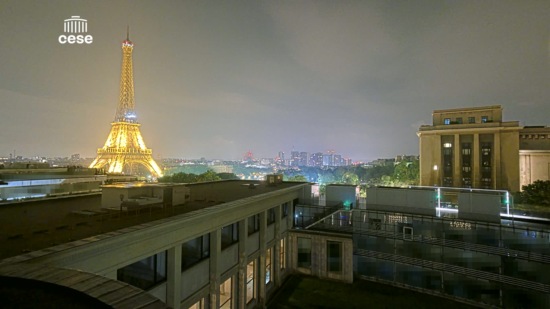 A nighttime view of the Eiffel Tower illuminated, with a city skyline in the background under a hazy sky, seen from atop a building.