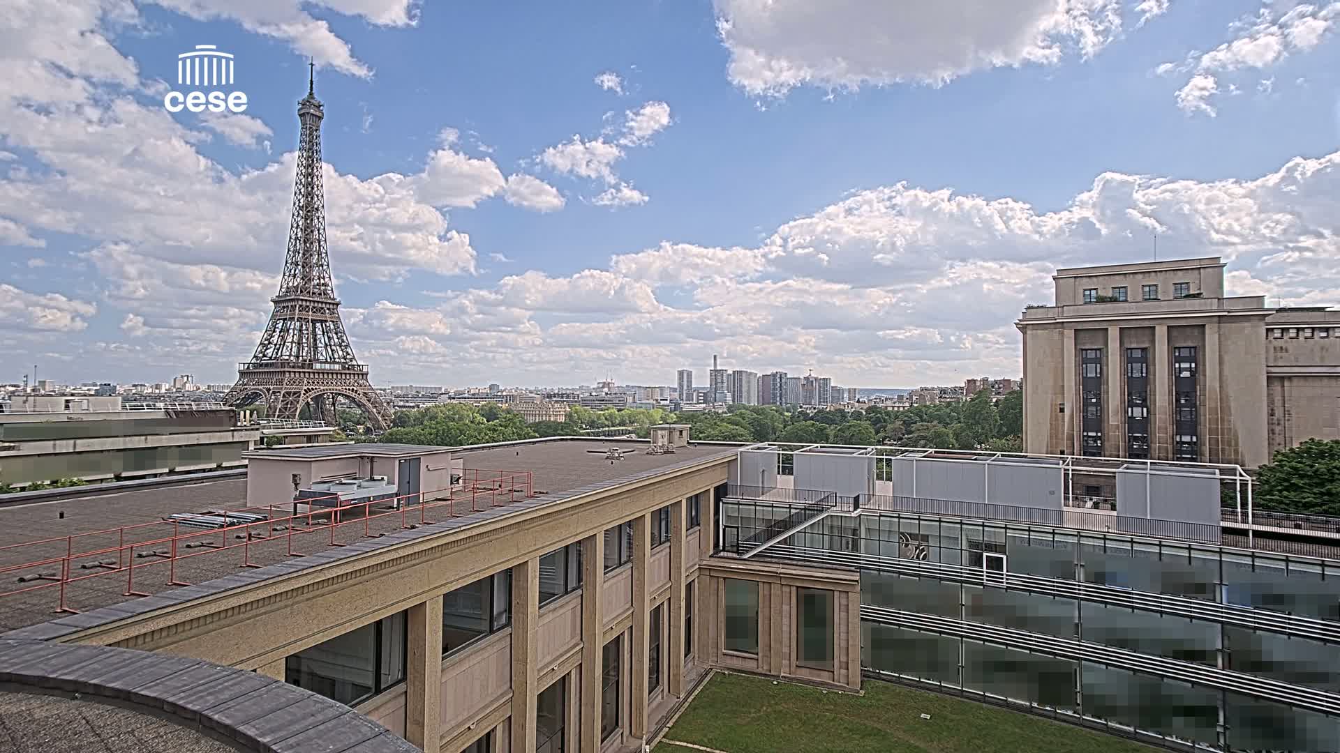 A view from a rooftop shows the Eiffel Tower in the distance under a partly cloudy sky, with other buildings and a grassy courtyard in the foreground.