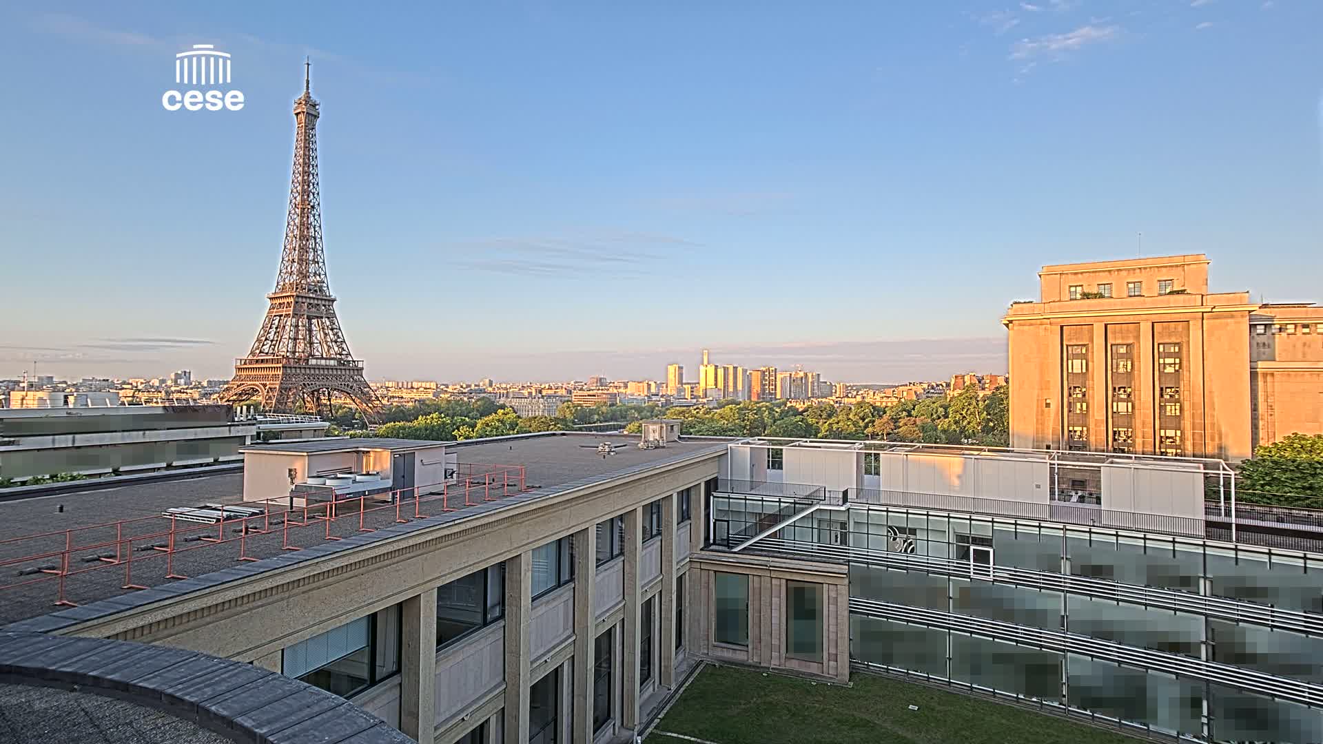 A view from a rooftop shows the Eiffel Tower in the distance under a clear, sunny sky, with other buildings and a cityscape in the mid-ground.