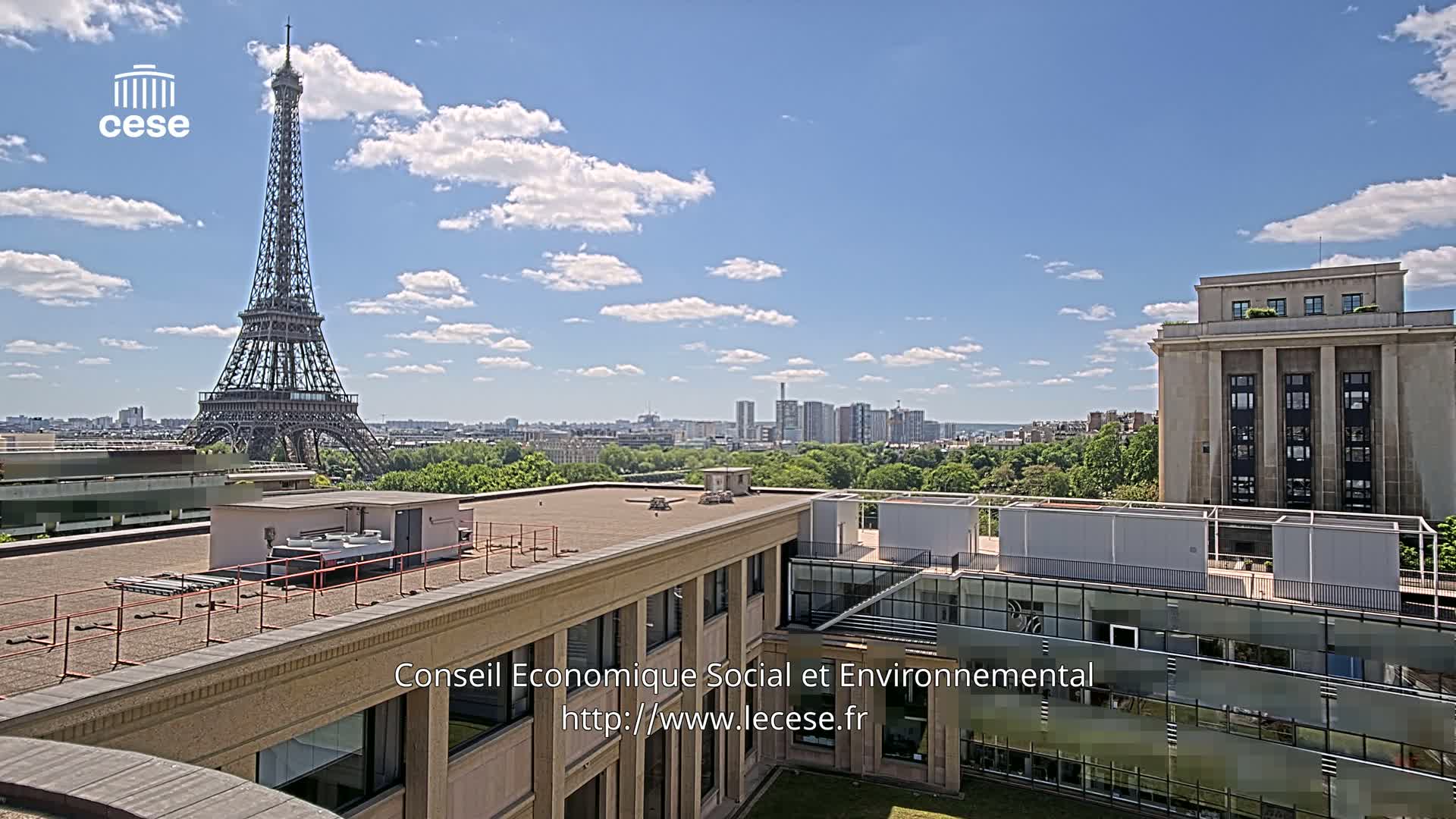 A rooftop view shows the Eiffel Tower in the distance under a partly cloudy blue sky, with a cityscape and green trees in the middle ground, and a large building to the right.