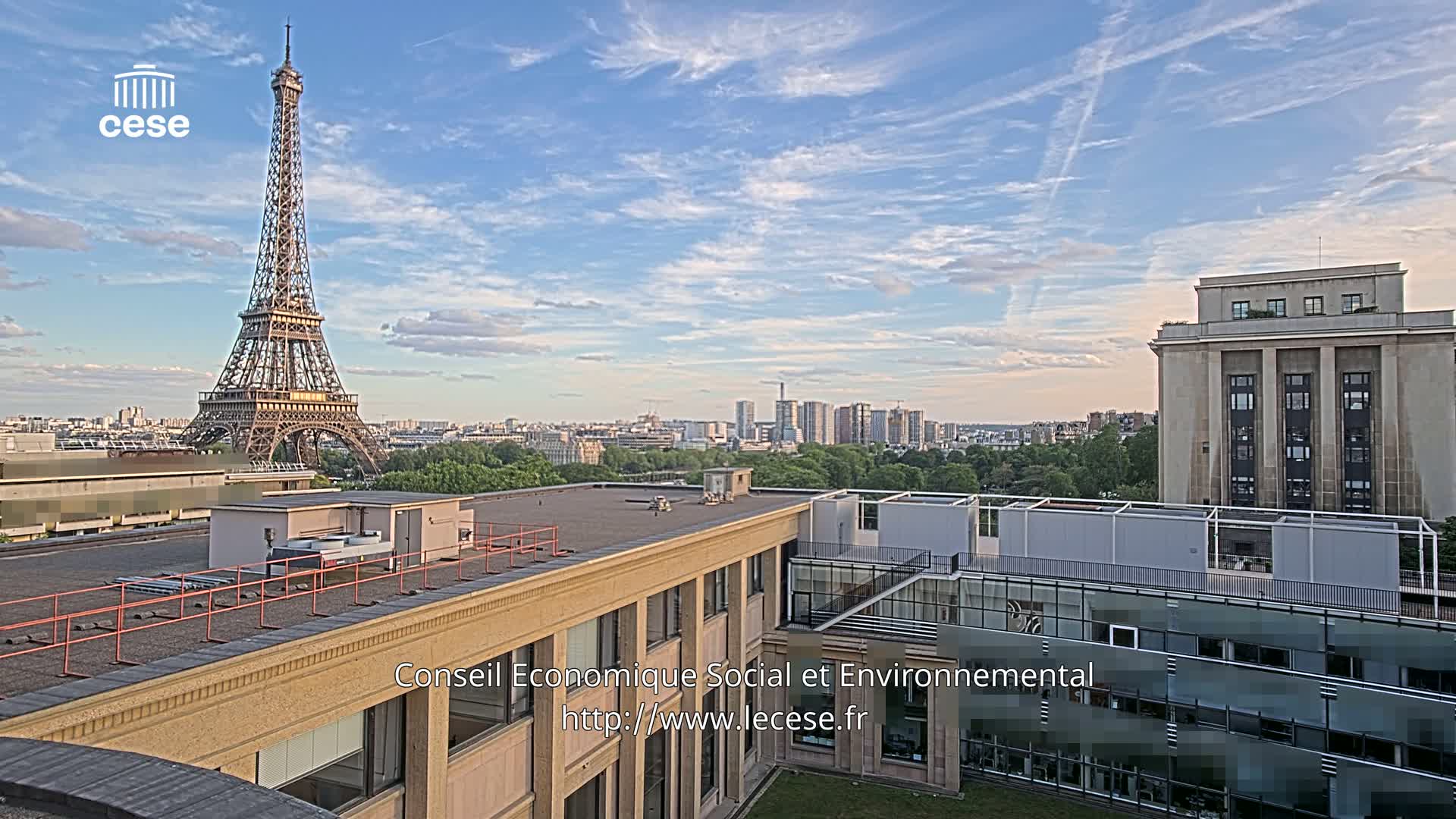 A rooftop view of Paris, under a mostly clear sky with a few wispy clouds, shows the Eiffel Tower in the distance.