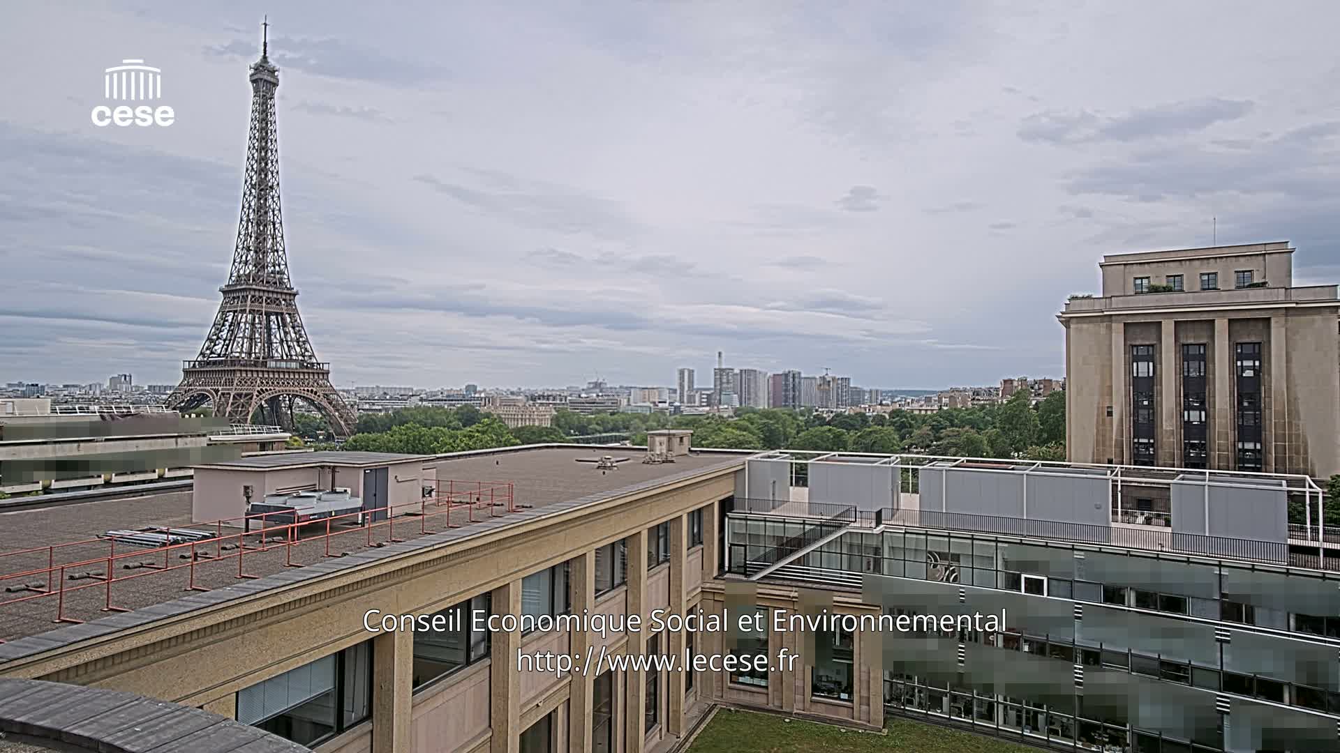 An overcast day view from a rooftop shows the Eiffel Tower in the distance, beyond a cityscape and green trees.