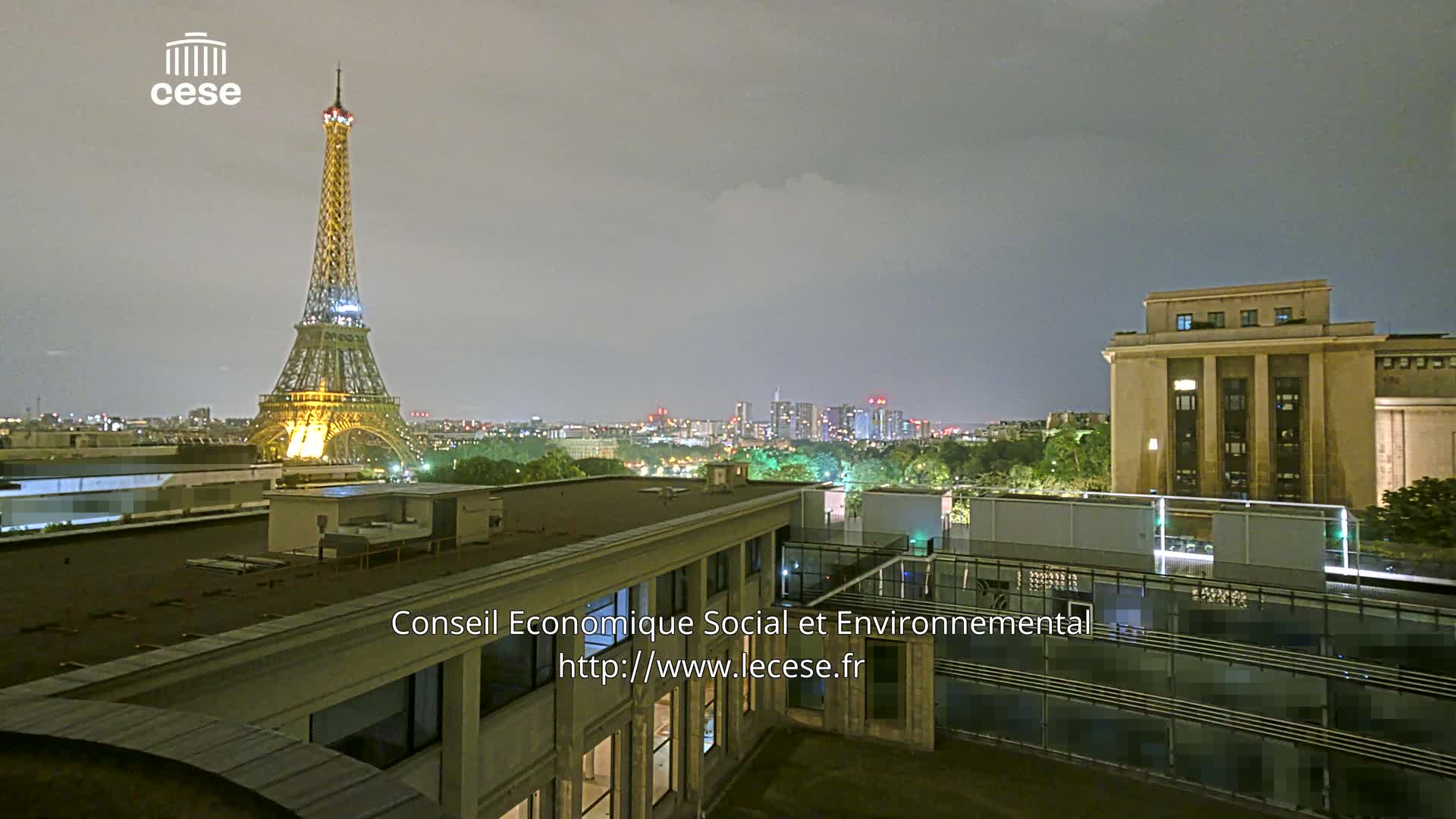 A nighttime view of the Eiffel Tower illuminated, a city skyline, and a large building, under a cloudy sky.