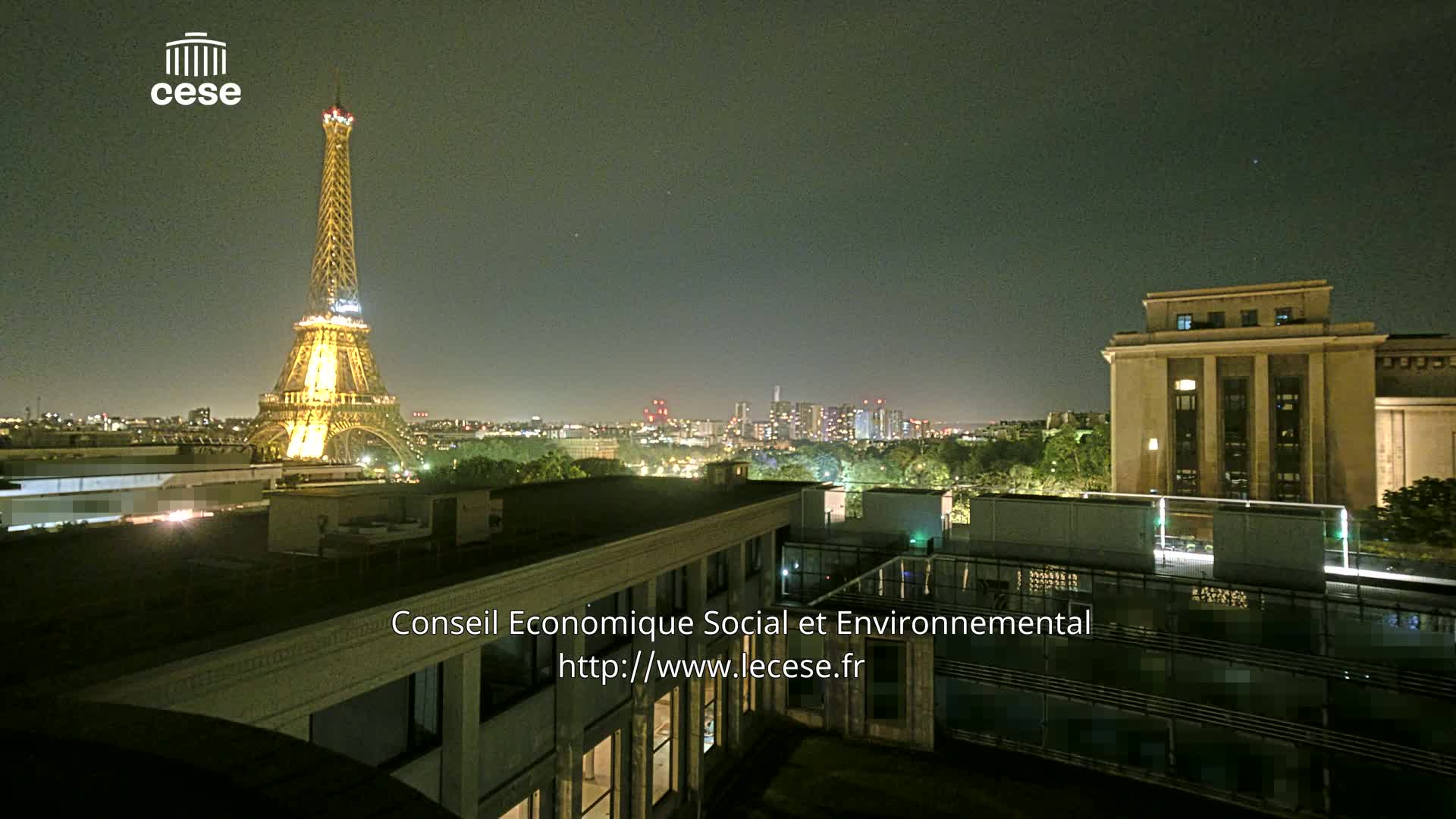 A nighttime view from a rooftop shows the Eiffel Tower illuminated, a cityscape in the distance, and a large building nearby.