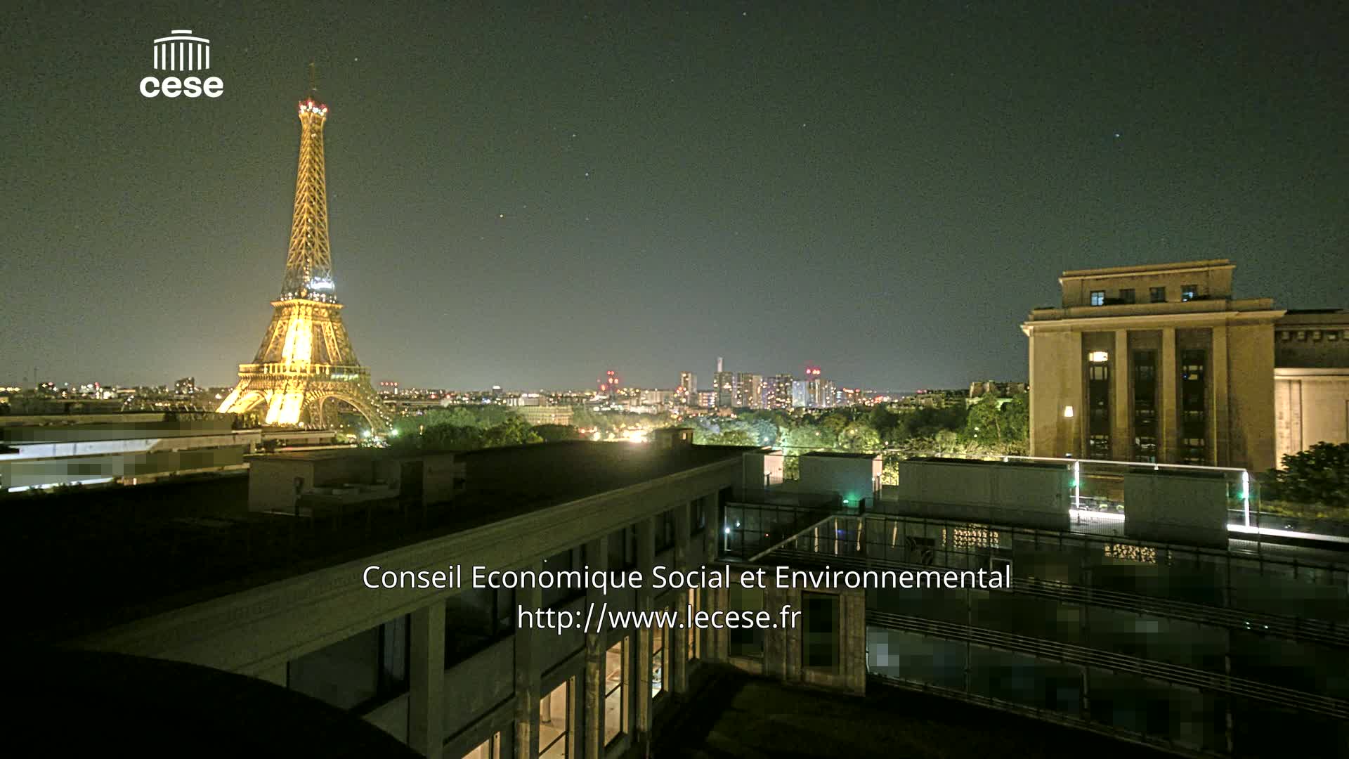 A nighttime view of the Eiffel Tower illuminated against a dark sky, with city lights and a large building visible in the foreground.