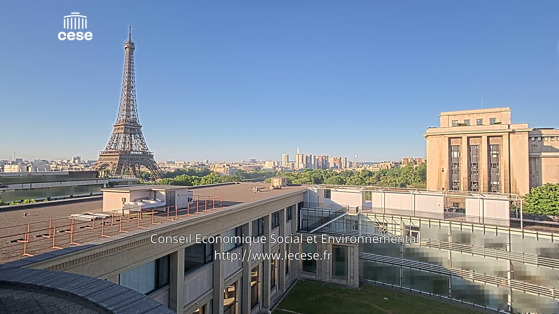 A rooftop view of a city on a clear, sunny day shows the Eiffel Tower in the distance and a large building in the foreground.